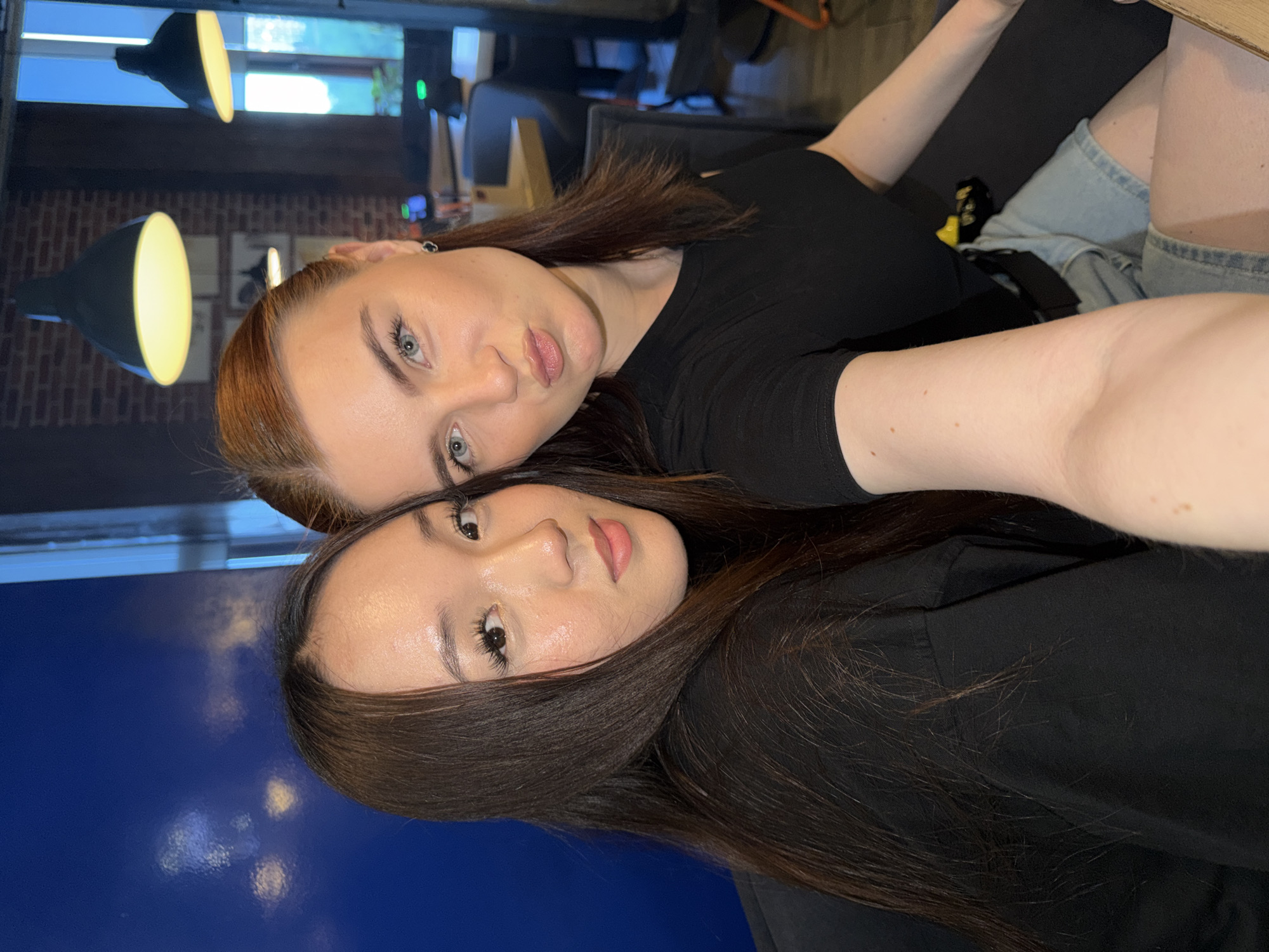The image displays two young adult females indoors in an establishment in Sinyavino, Russia, likely a cafe or restaurant, captured in what appears to be a selfie. The female on the left has long, straight dark brown hair with a middle part, dark brown eyes with elongated eyelashes, defined eyebrows, and light skin exhibiting a subtle sheen on her forehead, nose, and chin. She wears a black short-sleeved top and maintains a neutral expression with a slight smile while looking directly at the camera. The female on the right has reddish-brown hair, light blue-grey eyes, defined eyebrows, and light skin. She wears a black short-sleeved top and light-wash denim shorts. A small black stud earring is present on her left ear. She looks directly at the camera with a "duck face" expression. Her right arm, showing light hair and several small moles, is extended towards the camera, consistent with holding a mobile device. A dark-colored pouch or bag with a yellow strap/buckle is visible attached to her waist.
The background on the left consists of a solid dark blue wall with blurred circular light reflections. The background on the right reveals elements of an indoor commercial space, including an exposed red brick wall, a large window displaying a bright outdoor scene, and several industrial-style black pendant lamps emitting warm yellow light. Blurred furniture, including dark chairs and light-colored tabletops, along with a computer monitor displaying a green light, are visible further in the background.