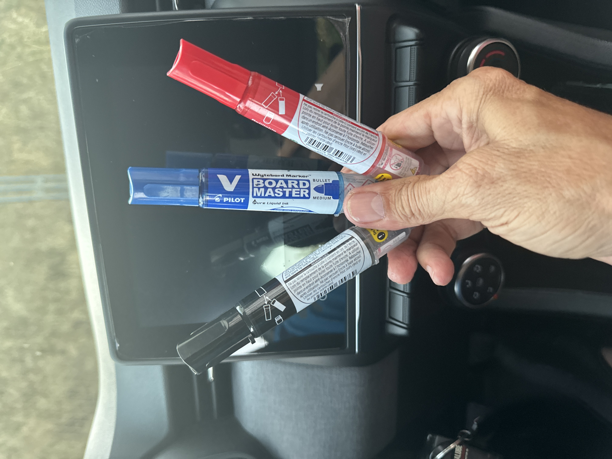 A hand holds three whiteboard markers in front of a car's digital display. The markers are black, blue, and red, and they are all Pilot Board Master brand. The car appears to be parked, possibly in an outdoor setting. The background is blurred, but suggests a natural environment with some foliage.
