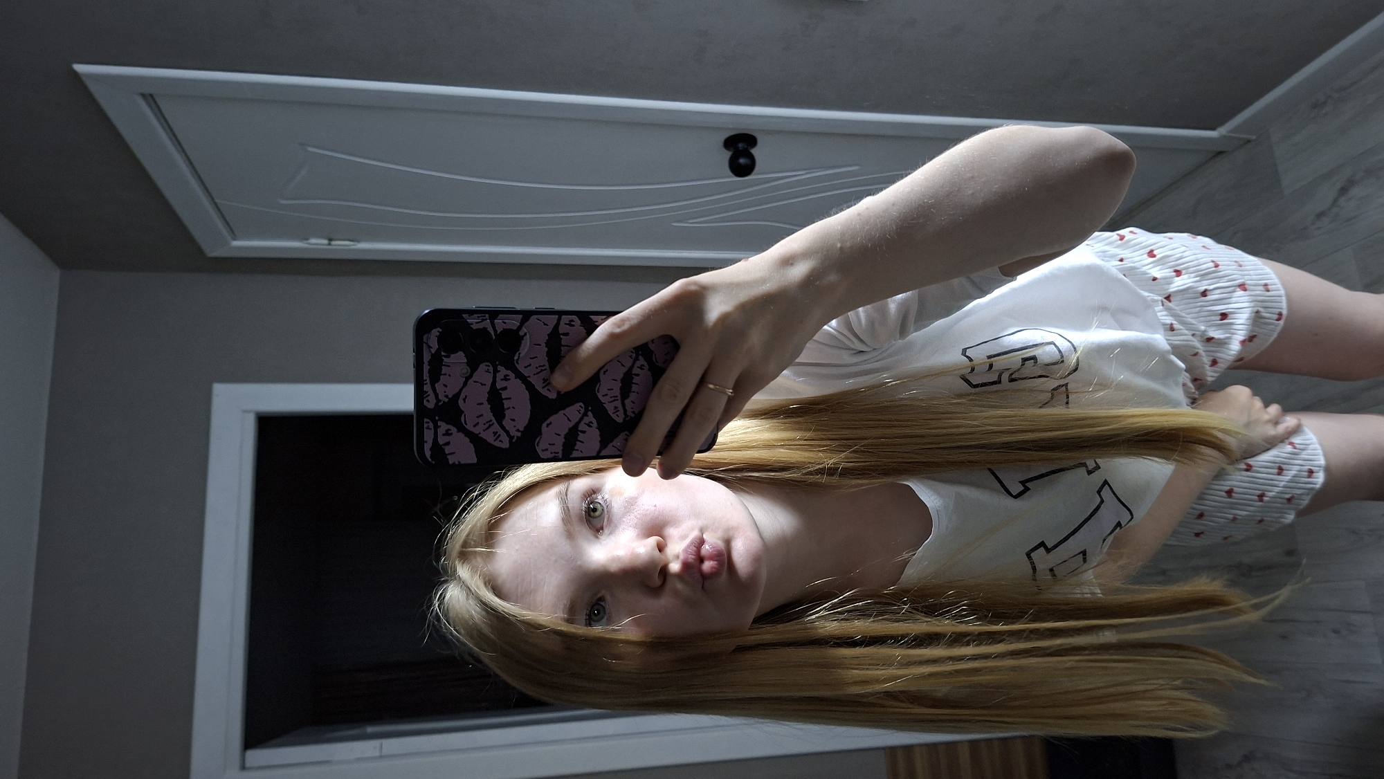 A young Caucasian woman with long blonde hair is taking a selfie in a room. She is wearing a white t-shirt with a black graphic and white shorts with a red heart pattern. Her lips are pursed for a kiss, and she is holding a smartphone with a purple case decorated with lips. A ring is visible on her right hand. The background shows a doorway with a white door and a textured grey wall. The floor appears to be wood laminate. The overall impression is a casual, indoor setting. The image does not contain any identifiable landmarks or information that would place it specifically in Chaikovsky, Russia.