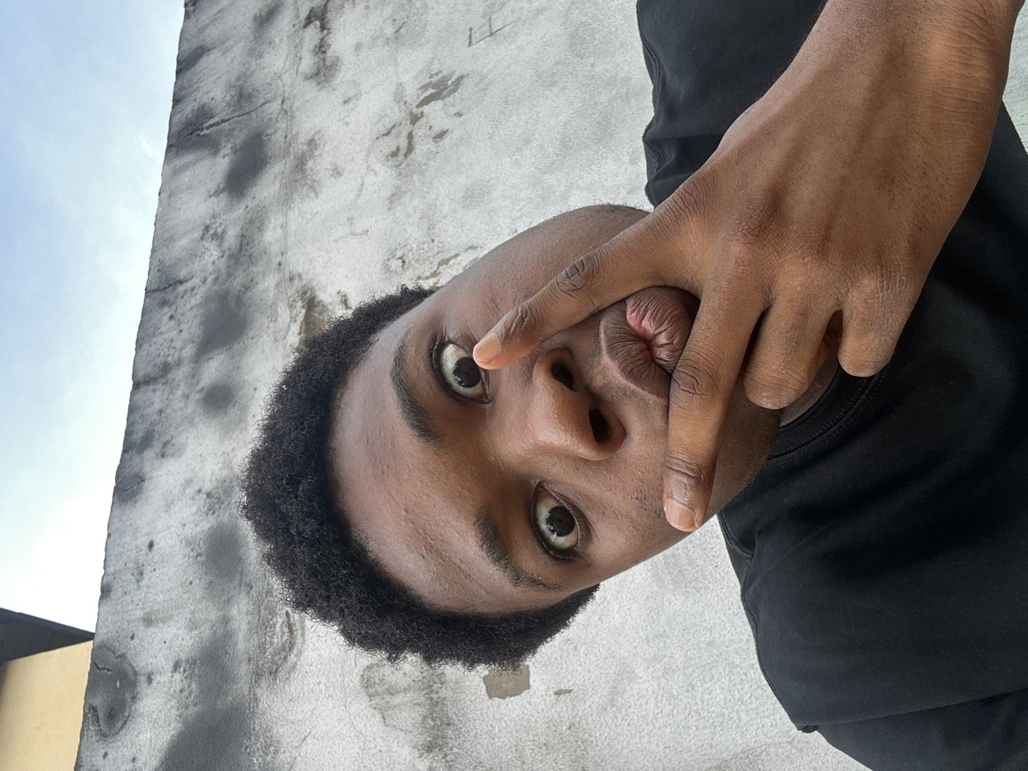 A medium shot captures a young Black male outdoors, his head tilted slightly upward and his body facing slightly left. He wears a black t-shirt. His left hand is raised to his face, with his index finger pressed against his lips, which are pursed. His right eye is wide open and looking directly at the camera, while his left eye is partially obscured by his index finger and the angle of his head. The background consists of a light-colored concrete wall with dark, irregular smudges and a small section of a yellow-painted structure at the top left. Above the wall, a pale blue sky with some clouds is visible. The setting appears to be an urban environment in Ihiagwa, Nigeria, suggested by the architectural style of the wall and building, and the ambient light.