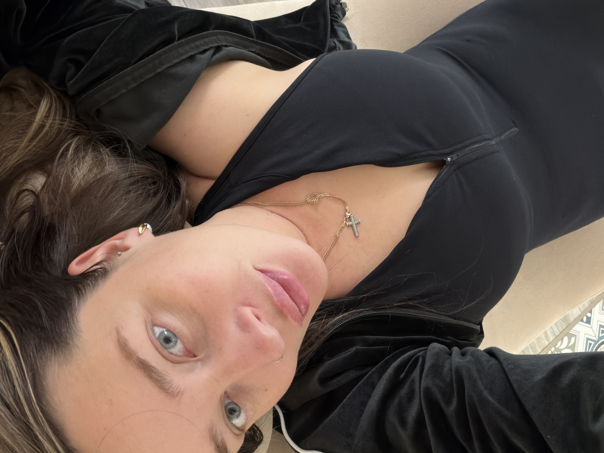 Here is a detailed description of the image:
A woman lies on her back, looking up at the camera. She has fair skin, blue eyes, and light brown hair. She wears a black, tight-fitting bodysuit with a zipper slightly open at the chest, revealing a gold necklace with a cross pendant. A black velvet jacket is partially visible, draped over her shoulders. She has piercings in her ear and nose. The background consists of a beige surface, presumably a couch or bed, and a glimpse of patterned flooring. The location is unspecified and unknown.