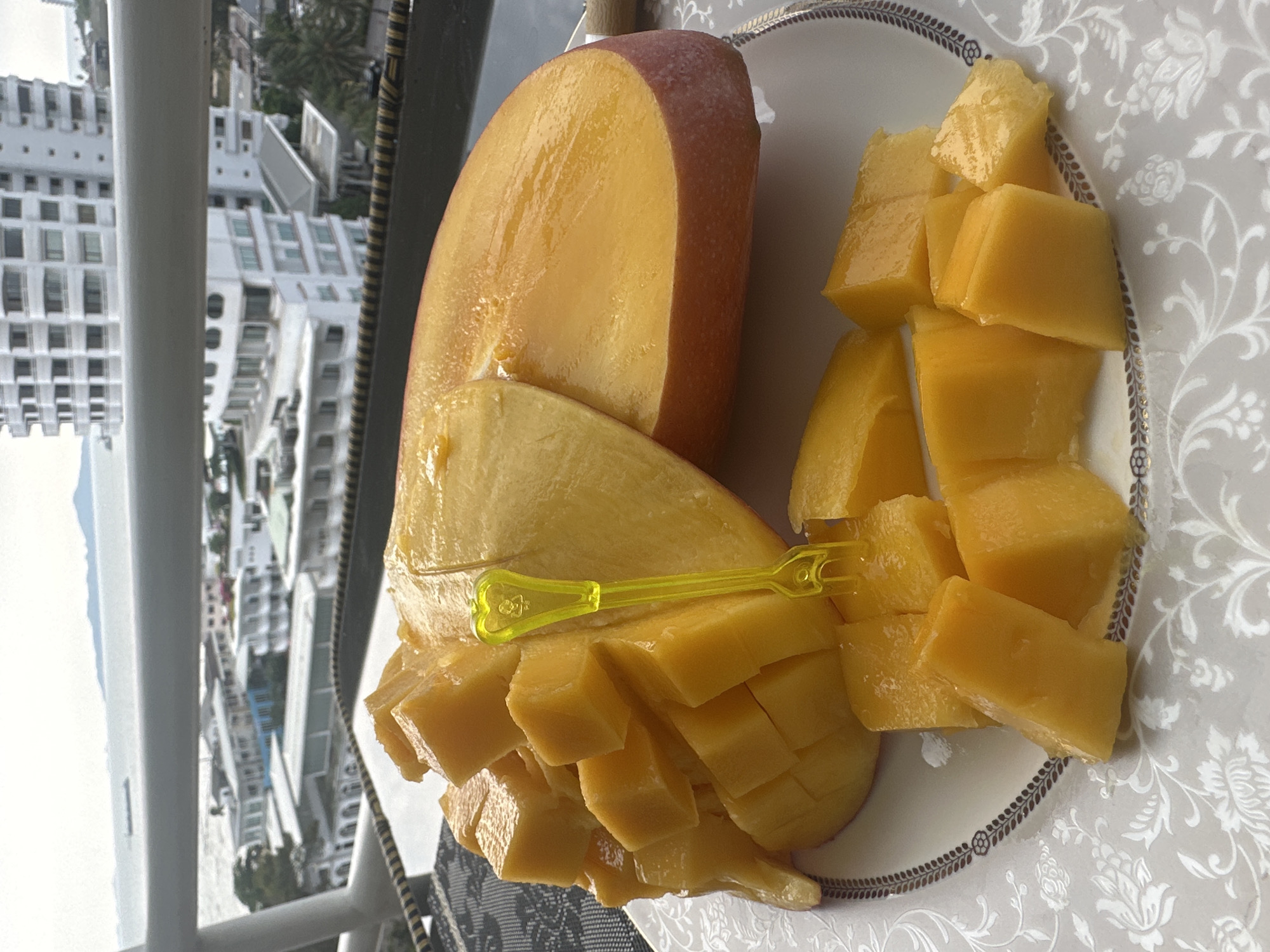 A partially-eaten mango, cubed and sliced, rests on an ornate plate. A small, yellow plastic fork pierces the fruit. The plate sits on a tablecloth patterned with white flowers on a gray background. Outside, a city scene is visible, featuring white high-rise buildings, likely in Pattaya City, Thailand. A railing is visible, suggesting the photo was taken from a balcony.
