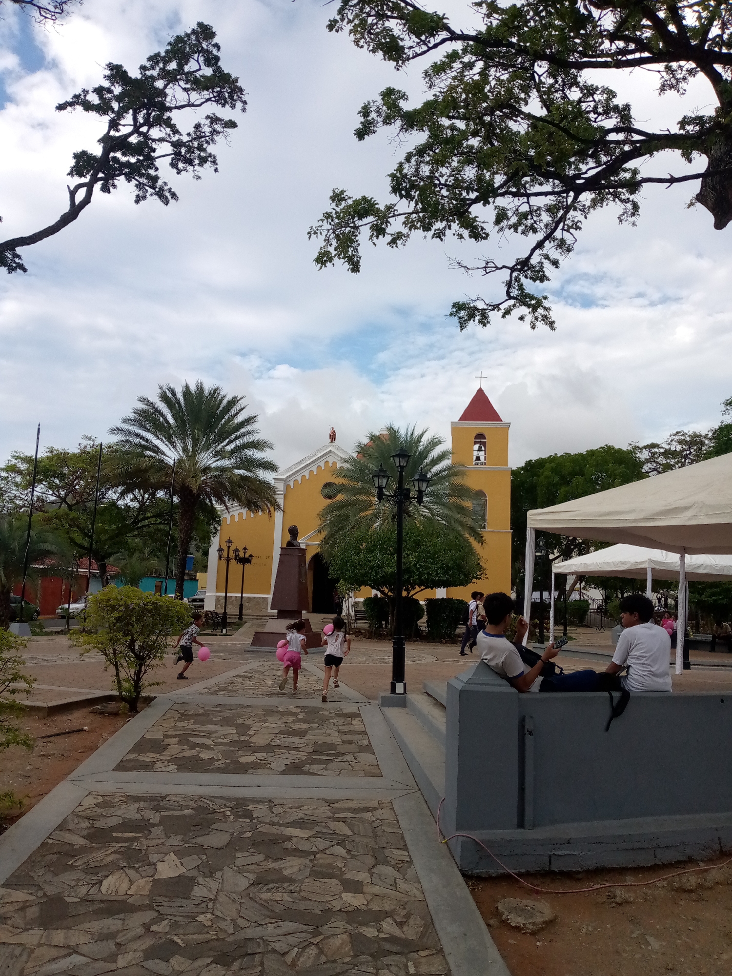 The image captures a sunny day scene in the city of San Juan Bautista, Venezuela, featuring a yellow church with a red-roofed bell tower. In the foreground, a stone pathway leads towards the church, with several children running on it, some holding pink balloons. To the right, two young people are seated on a gray concrete structure, one looking at a phone and the other engaged in conversation. Palm trees and other lush green foliage surround the church and the surrounding plaza, adding to the tropical ambiance. A white tent, possibly for an event, is visible in the background, providing shade. The sky is partly cloudy, with patches of blue sky visible.