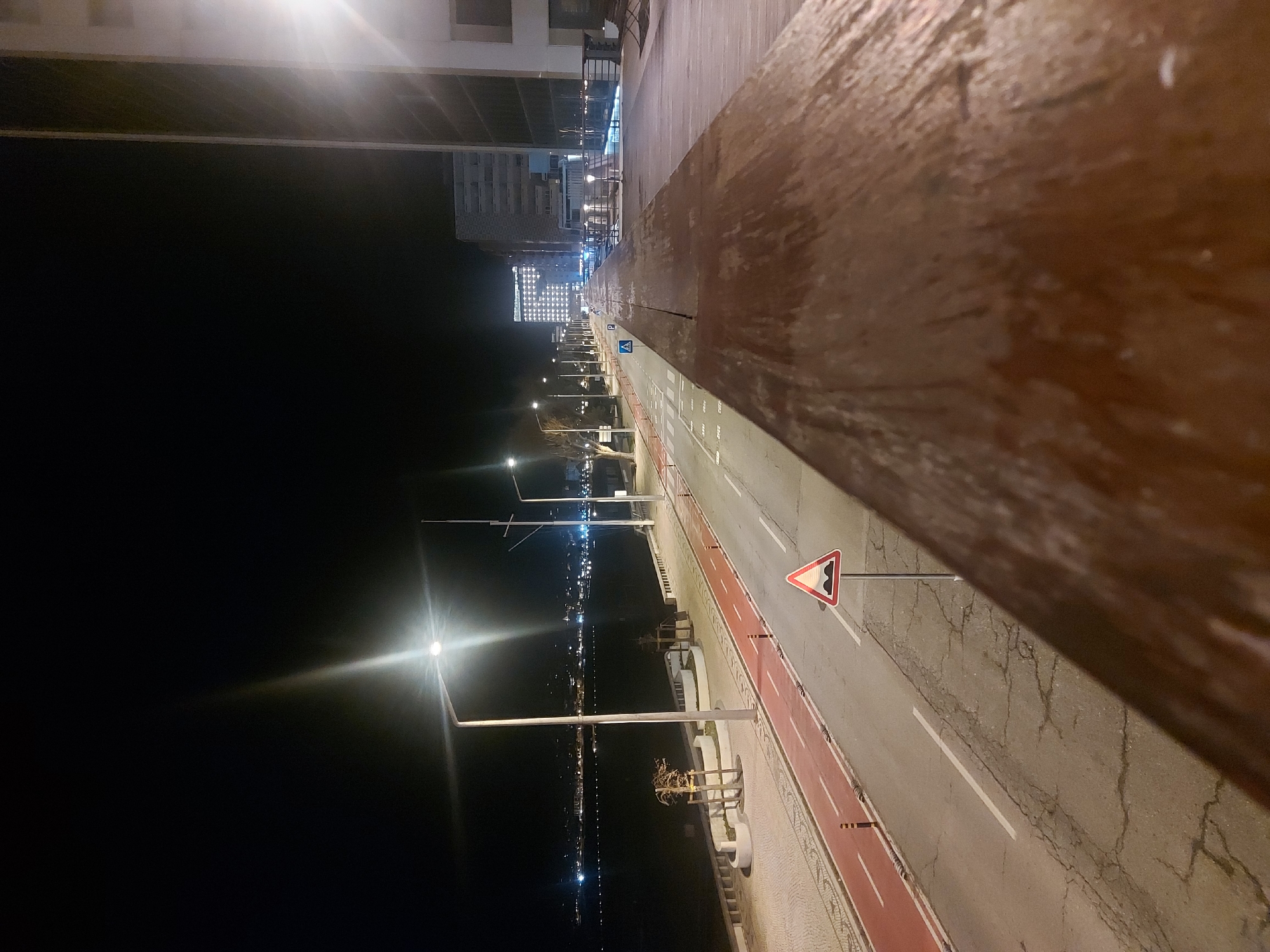 The image shows an elevated view of a street in Figueira da Foz, Portugal, at night. A wooden railing is visible in the foreground. A street with a designated bike lane is illuminated by streetlights. A road sign is also visible along the side of the road. In the distance, buildings with illuminated facades can be seen. The image captures a dark sky and the glow of city lights reflecting on the water.