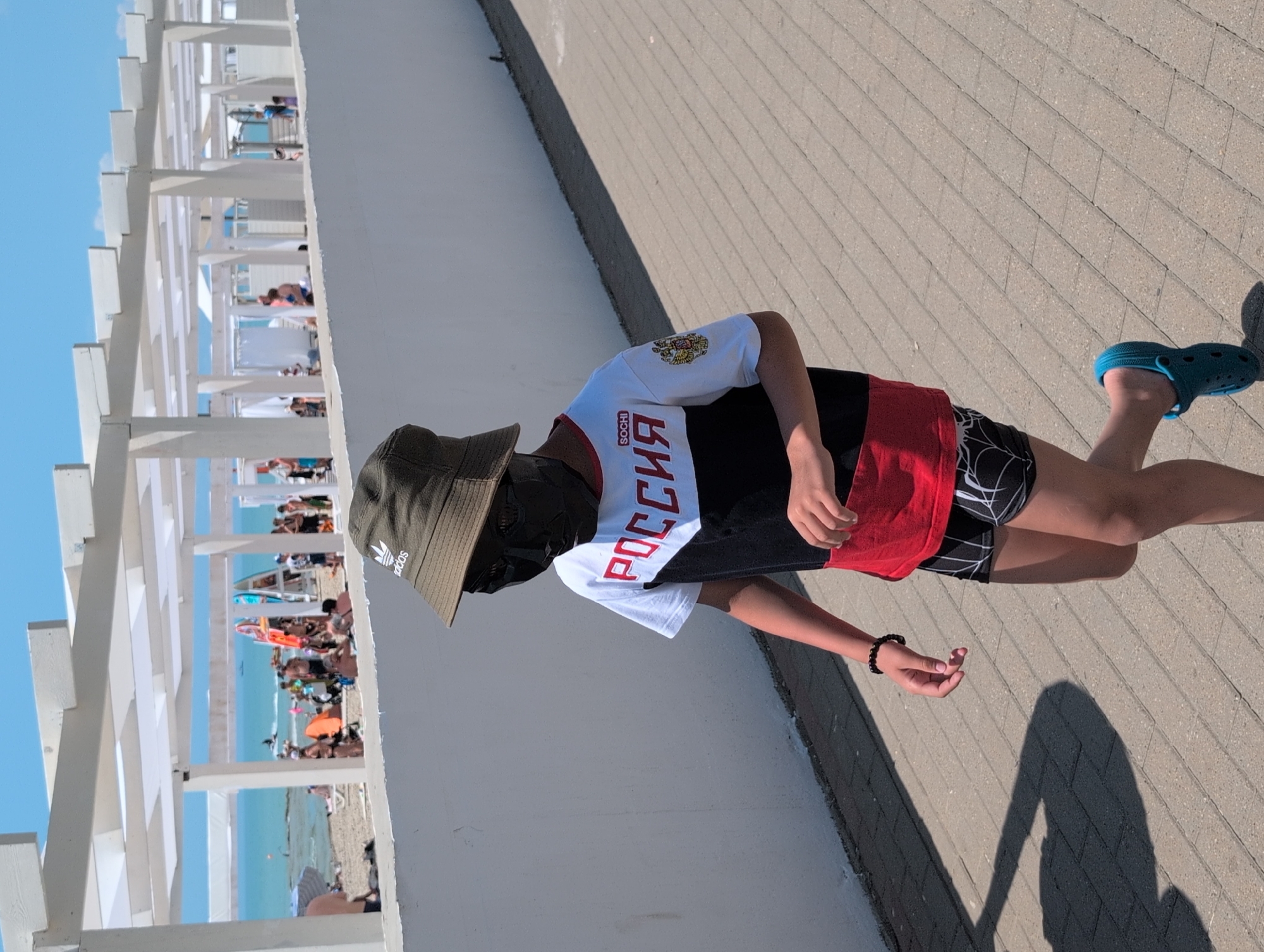 A young person wearing a dark mask, a bucket hat, a t-shirt with red, black, and white stripes and "РОССИЯ" and "SOCHI" written on it, and black shorts with a white pattern, walks on a paved walkway. The person is wearing blue clogs. In the background, there is a beach with people, umbrellas, and beach structures. The sky is blue and clear, and there is a body of water that appears to be the Black Sea. This scene is likely in Divnomorskoye, Russia, a coastal resort town.