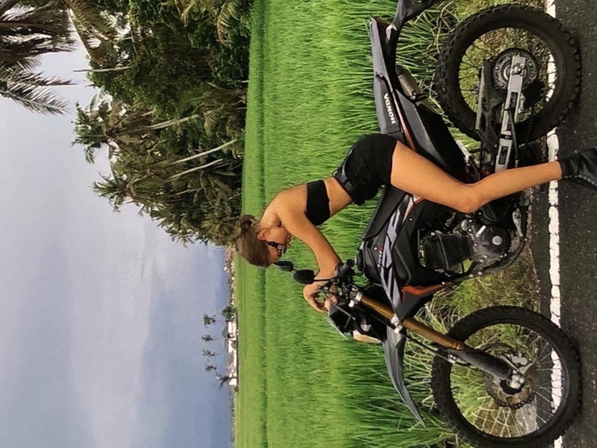 A young woman with her hair pulled back is kneeling on a black dirt bike with gold forks, in front of a lush green rice paddy field. She is wearing a black bandeau top and black shorts. The background shows palm trees and a cloudy sky. The dirt bike has "HONDA" written on the rear fender. The scene is set in a rural landscape, possibly in Southeast Asia, not in Bratislava, Slovakia.