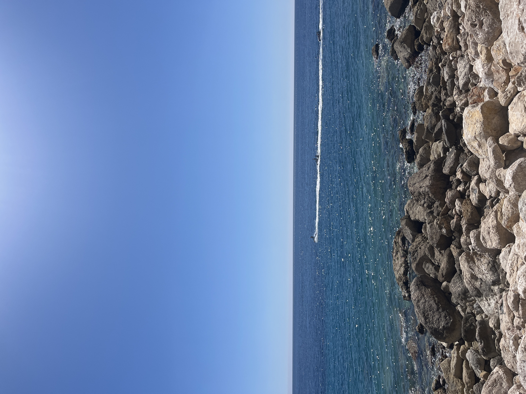 The image shows a coastal scene in El Poble Nou de Benitatxell / Benitachell, Spain. The sky is clear and blue, occupying the left side of the frame. To the right, a body of water stretches out, with a trail of white water suggesting a boat or jet ski is moving across it. Along the edge of the water, a rocky shoreline is visible. The rocks are various shades of gray and brown, creating a rugged texture against the water's surface.