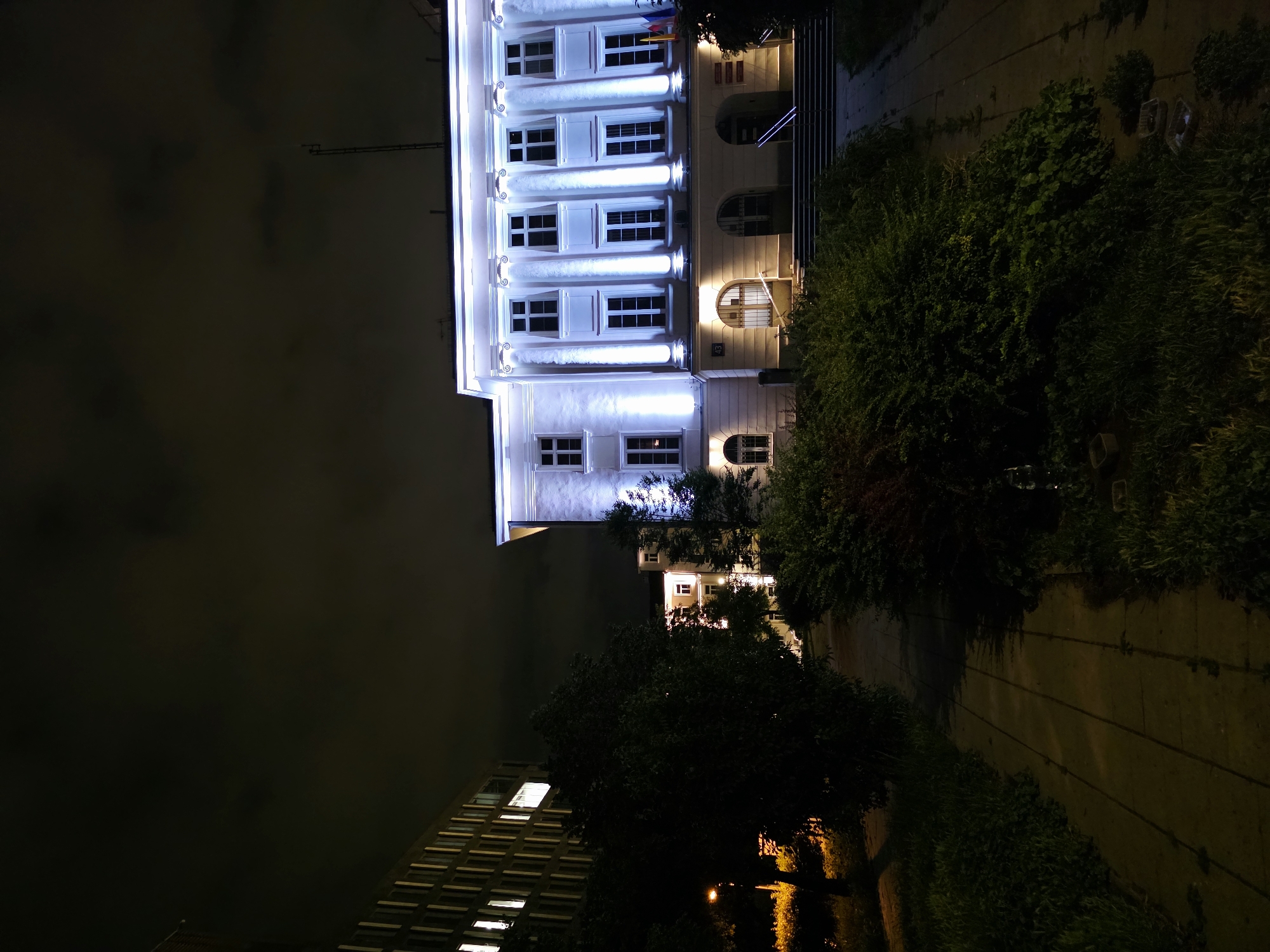 The image shows a night view of a well-lit building with classical architectural elements, possibly a public institution, located in Warsaw, Poland. The building features a white facade adorned with columns and numerous windows, illuminated by bright blue lights. In front of the building, there is a set of stairs leading up to the entrance, with handrails. Adjacent to the main building, a taller, modern building with rows of illuminated windows is visible to the left. The foreground consists of a pathway bordered by lush green bushes and trees, suggesting a park or a well-maintained public space. The sky above is dark and cloudy.