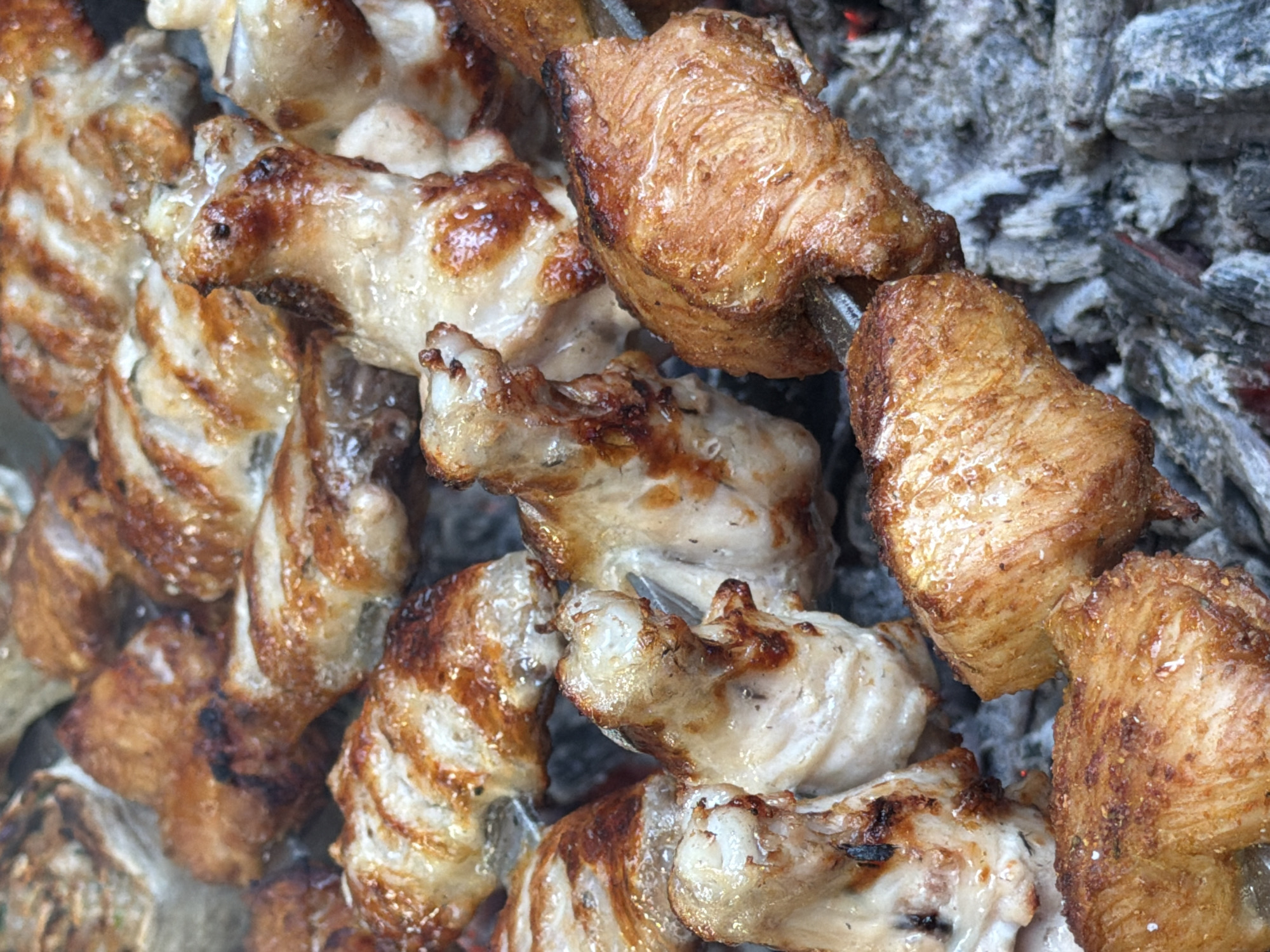 A close-up, overhead view shows grilled chicken wings on a skewer and resting on hot coals. The chicken wings are golden brown with some charred edges. The coals are gray and appear to be glowing red in places. The background is out of focus but suggests more food items. The scene depicts food being prepared outdoors. There is no specific indication of the location being Tuapse, Russia.