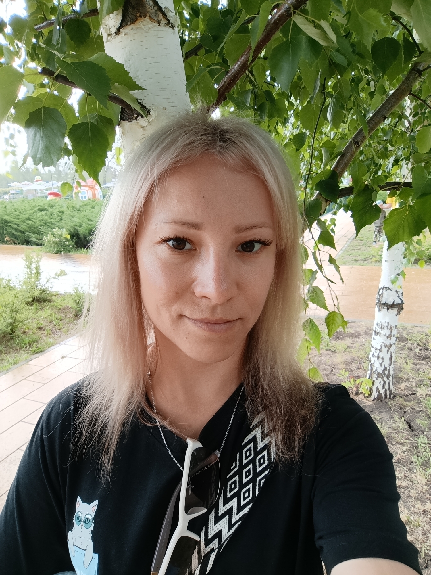 A selfie is taken of a young Caucasian woman with blonde hair in a park. She is wearing a black t-shirt with a graphic of a cat wearing sunglasses, and a necklace. She is outdoors, with birch trees and green leaves in the foreground. Behind her, there is a path, some grass, and a body of water. In the distance, there are some colorful structures that appear to be part of an amusement park, which might indicate the location as "Novokuybyshevsk Park" in the city of Novokuybyshevsk, Russia. The sky is overcast, suggesting a cloudy day.