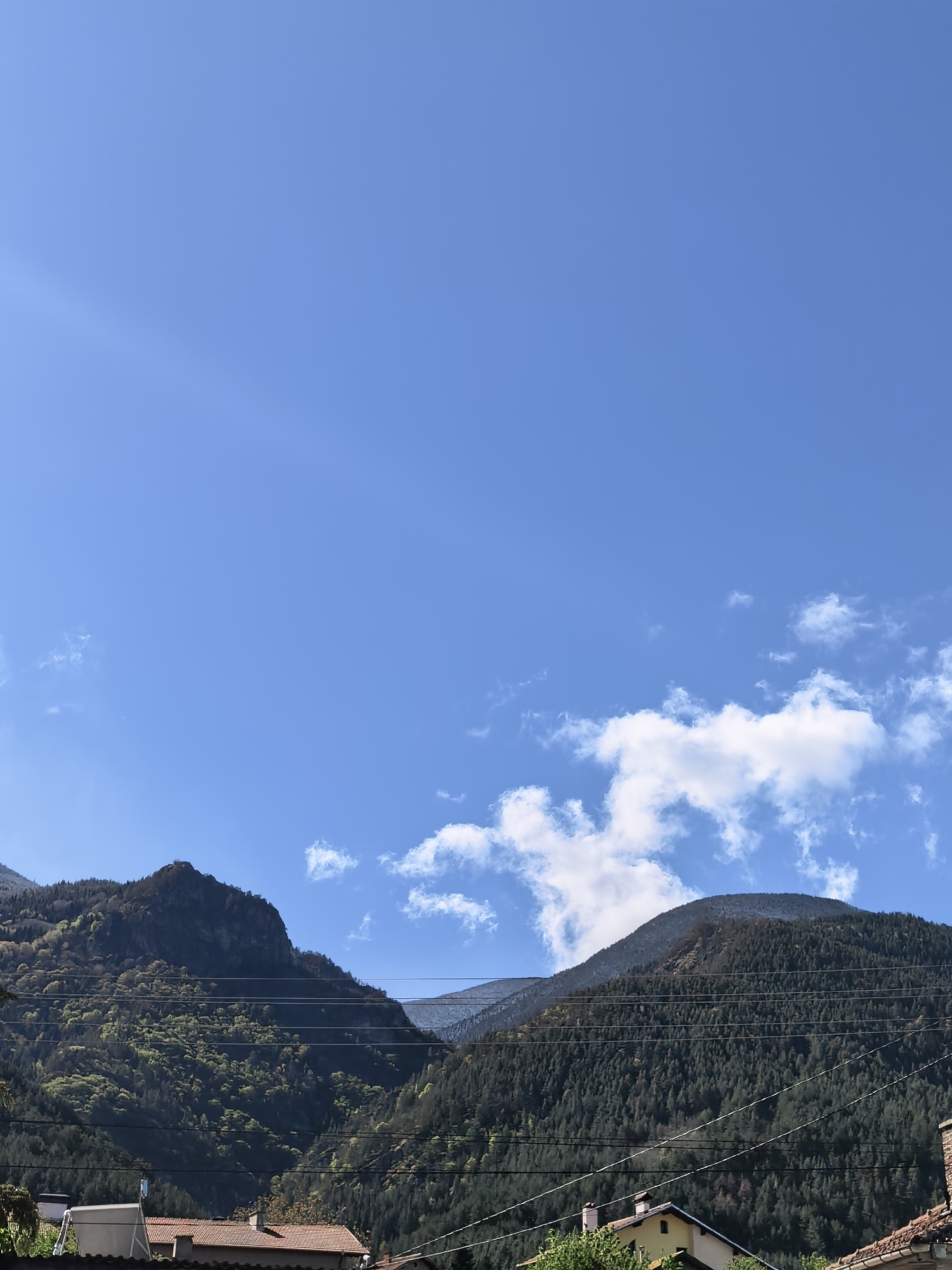 The image depicts a view of mountainous terrain under a clear blue sky with scattered white clouds. The mountains are covered in dense forestation, with some patches of green vegetation interspersed among the darker trees. Power lines are visible running across the mountains in the foreground. The rooftops of houses can be seen at the bottom of the frame, indicating a populated area.
Based on the provided information, the location is identified as Ovchartsi, Bulgaria.