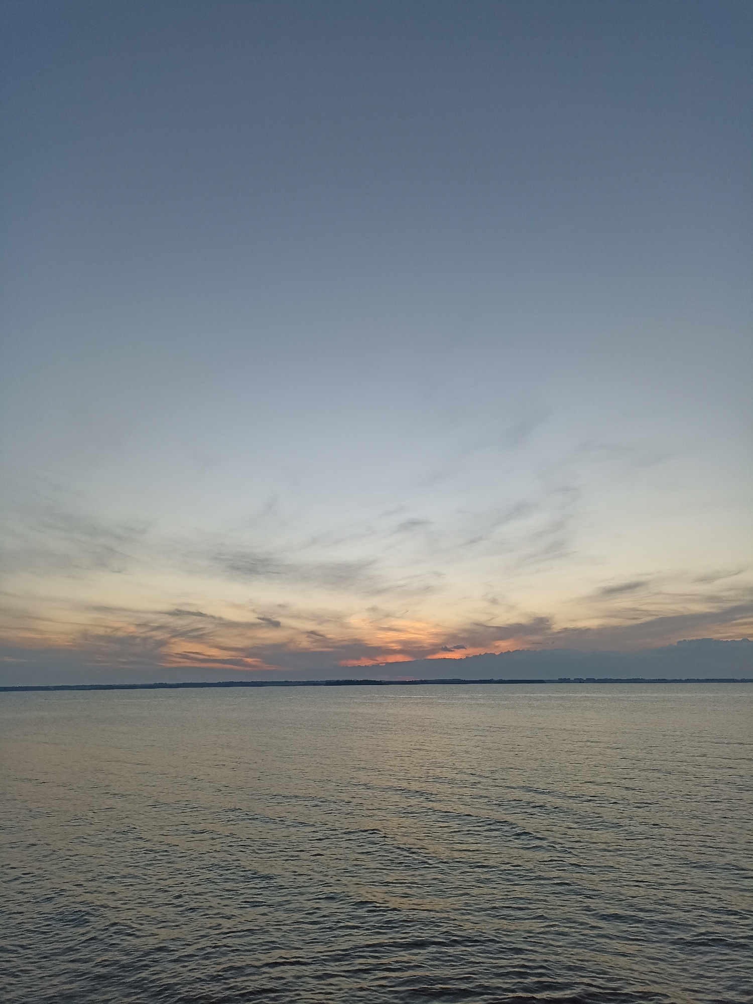 The image depicts a serene sunset over a body of water, likely a lake or river, viewed from the shore of Zavyalovo, Russia. The sky is a gradient of soft blues and grays, transitioning to warm hues of orange and pink where the sun is setting on the horizon. Wispy clouds are scattered across the sky, catching the last light of the day. The water is calm, with gentle ripples reflecting the colors of the sky. A thin, dark line of land is visible in the distance, likely the opposite shore of the body of water or a distant cityscape. There are no people, animals, or man-made structures clearly identifiable in the image. The overall atmosphere is peaceful and tranquil, capturing a moment of natural beauty at dusk.