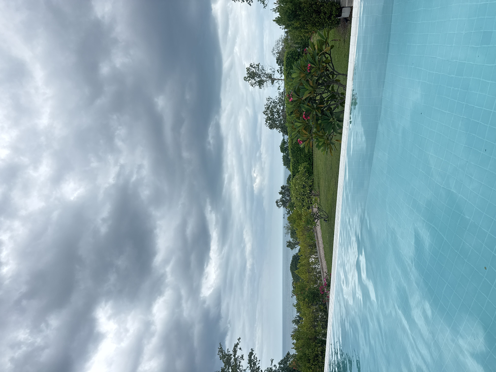 A high-angle, wide shot captures a view of a swimming pool and the ocean on a cloudy day. The foreground is dominated by the bright blue water of the infinity pool, with the tiled bottom visible beneath the surface. The middle ground features a lush green lawn, a pathway, and various trees and bushes, some with pink flowers. In the distance, the calm ocean stretches to the horizon, meeting a sky filled with dramatic, dark gray clouds. The scene is serene and suggests a tropical resort location in Ko Samui District, Thailand. There are no people visible in the image.