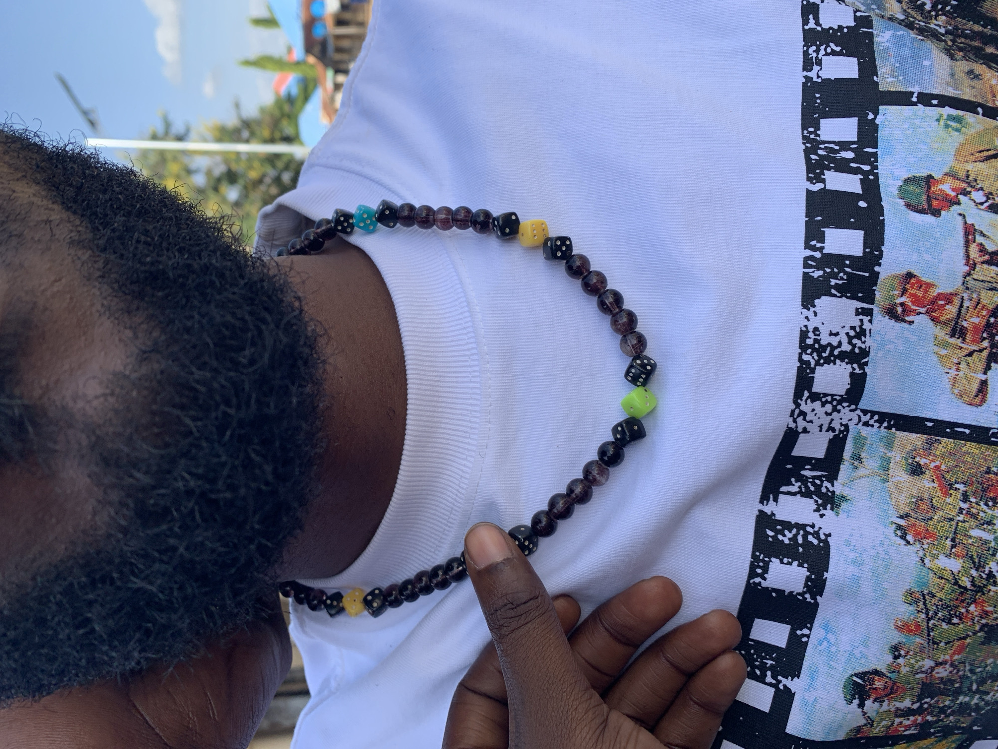 The image displays a close-up, chest-level view of an individual outdoors in New Stadium, Nigeria. The subject, exhibiting dark skin, possesses a dense, dark beard covering the lower facial area. A white, crew-neck t-shirt is worn, featuring a multi-frame film strip graphic across the chest depicting numerous soldiers in military helmets and uniforms, some holding rifles, against a light-colored background.
A beaded necklace is visible around the subject's neck, resting against the white t-shirt. The necklace consists of dark, translucent, spherical beads interspersed with square, dice-shaped beads. The dice are primarily black with white pips, accented by individual beads in yellow, light blue, and neon green. A dark-skinned hand, with the thumb and forefinger visible, is positioned on the left side of the frame, gently grasping the necklace at the center. The thumb's fingernail is visible and appears clean.
The background shows a clear blue sky with scattered white clouds in the upper right quadrant. A light-colored pole extends vertically in the background, behind the subject's right shoulder. Green foliage and trees are visible beneath and around the pole. Distant structures with visible red and blue roofs are present in the far right background.