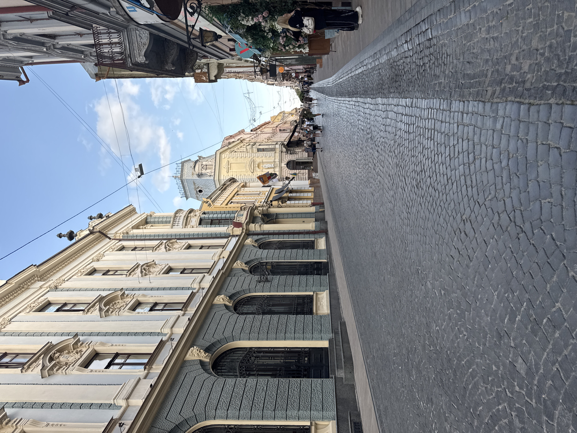 This is a low-angle, sideways shot of a cobblestone street in Chernivtsi, Ukraine. The street is lined with ornate, historical buildings with light-colored facades and dark window frames. The buildings feature intricate architectural details, including decorative carvings and arched windows.
Several people are walking down the street, appearing as small figures in the distance. Some are gathered near cafes or shops situated along the buildings. The scene suggests a leisurely day, likely in the late morning or afternoon given the bright sky and clear weather. The cobblestone street is well-maintained, with a slight sheen indicating it may have been recently wet or is simply reflecting the light.
Overhead, power lines crisscross the sky, typical of an urban setting. In the far distance, more buildings rise, some with prominent domes or spires, hinting at the historical and possibly religious significance of the area. The overall impression is one of an old European city, bustling with everyday life. No visible text is present in the image.