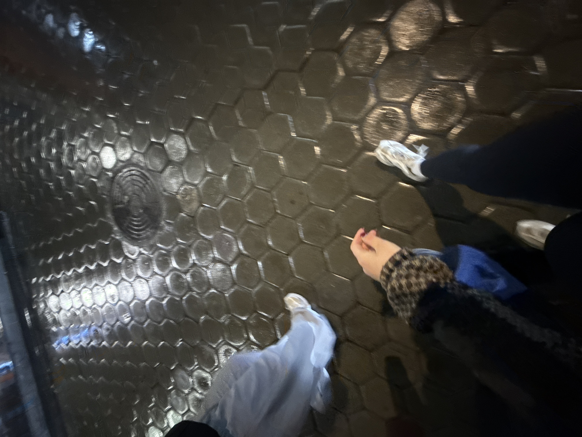 The image shows a ground-level view of a wet, hexagonal-tiled surface, likely a sidewalk. Light reflects off the wet tiles, giving them a shiny appearance. Three sets of feet are visible, indicating the presence of three people. One person wears white sneakers and black pants. Another person wears light-colored shoes and light-colored clothing. A third person wears silver sneakers and black pants. The arm of a person in a spotted jacket can be seen. One person is holding a pink item, probably a cigarette. The location is Yerevan, Armenia, as specified.