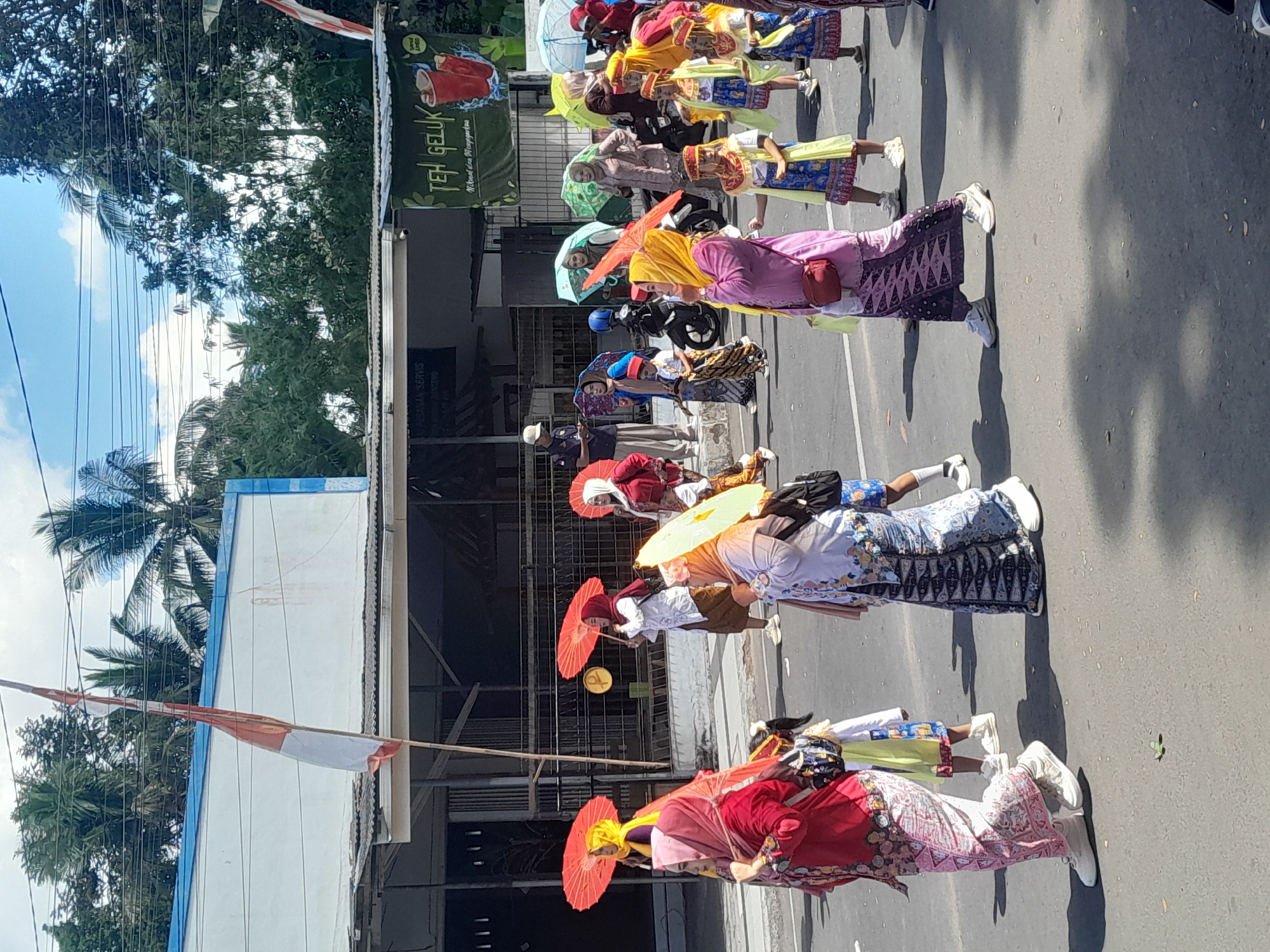 The image depicts a cultural parade taking place on a street in Genteng Kulon, Indonesia. The parade features several individuals, including women and children, dressed in traditional attire. Many participants are holding colorful umbrellas, predominantly red and yellow, likely for shade. The people are walking on a paved road, with buildings and trees lining the background. A banner with Indonesian text and an image of red drinks is visible on one of the buildings. The weather appears sunny with a clear sky and scattered clouds. A national flag of Indonesia is also prominently displayed, attached to a pole and partially visible against the sky and buildings. The scene suggests a festive and communal event, possibly a cultural celebration or festival.