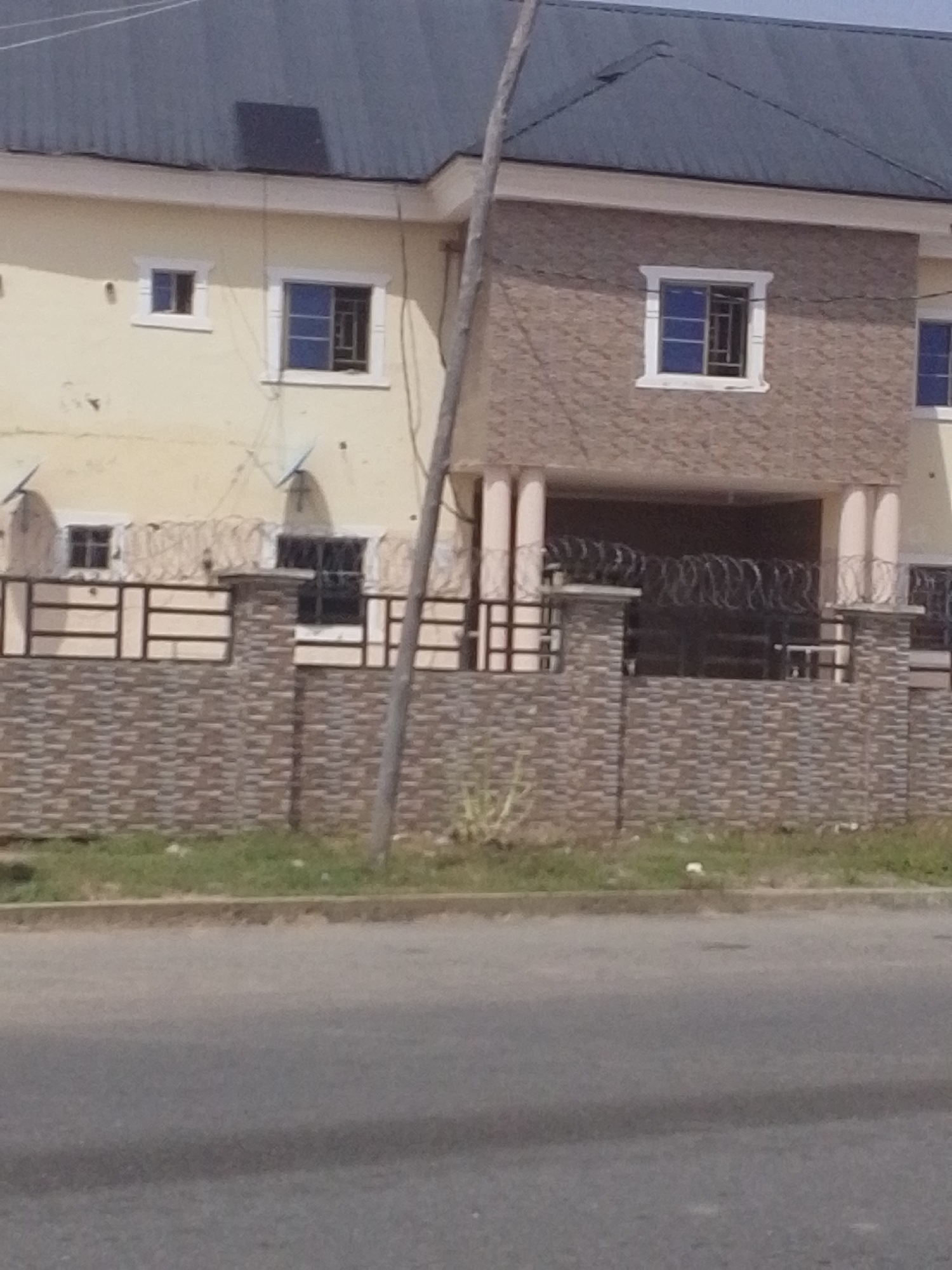 This image depicts a three-story building in Jalingo, Nigeria, viewed from a street. The building has a light yellow facade on the upper floors and a brick-patterned lower section with a covered entryway. There are several windows visible, some with blue panes and metal bars, and a dark-colored solar panel on the grey, corrugated metal roof. The building is bordered by a stone and metal fence topped with barbed wire. A utility pole stands in front of the building, and the street in the foreground is paved with asphalt. The environment appears to be urban or suburban, with some sparse green vegetation and debris along the roadside.