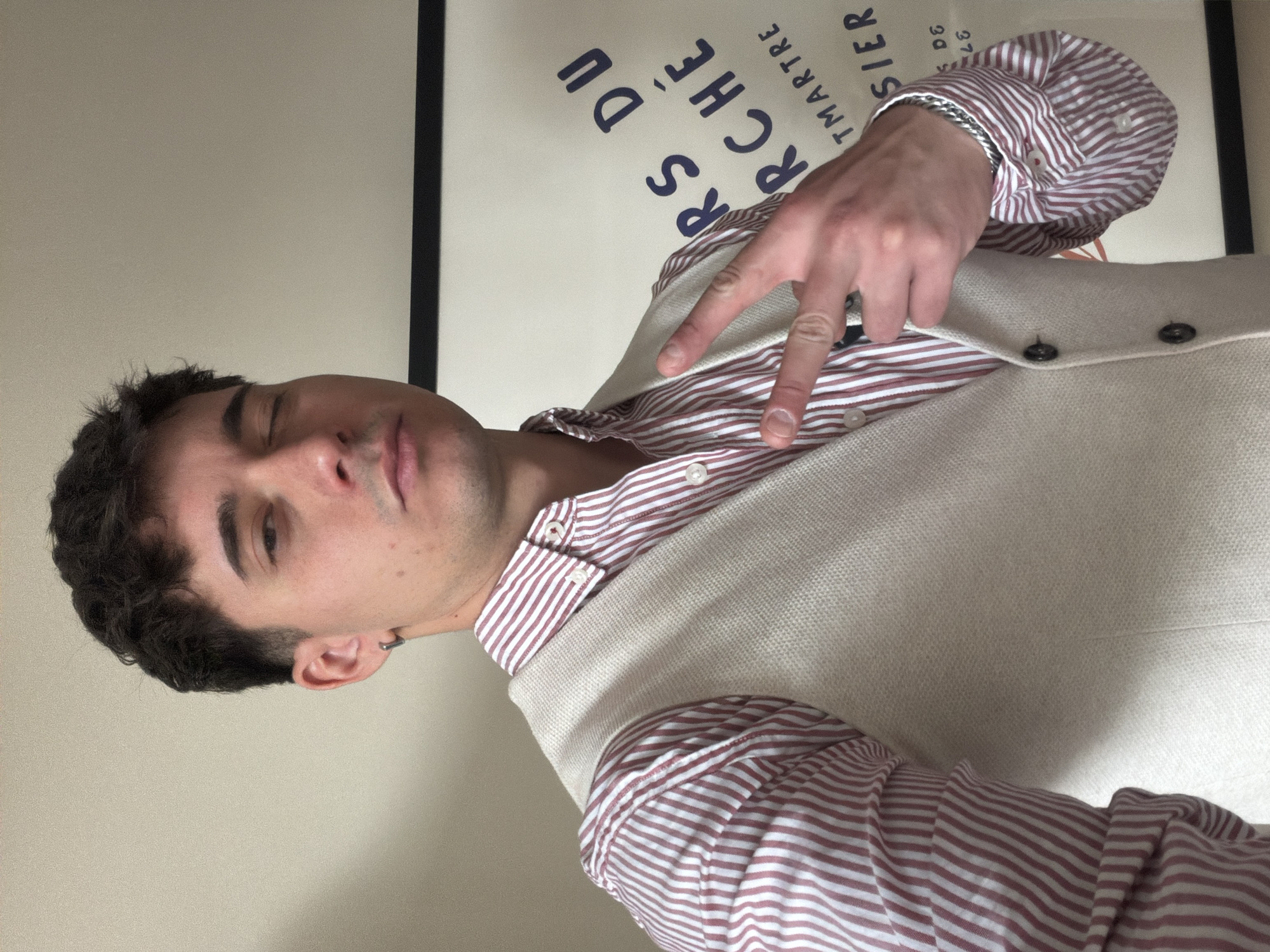 A man with dark hair and an earring is making a peace sign. He is wearing a striped shirt under a vest. Behind him is a framed print with the words "ARCHE' DU MARTRE" on it. No information suggests the location is Sofia, Bulgaria.