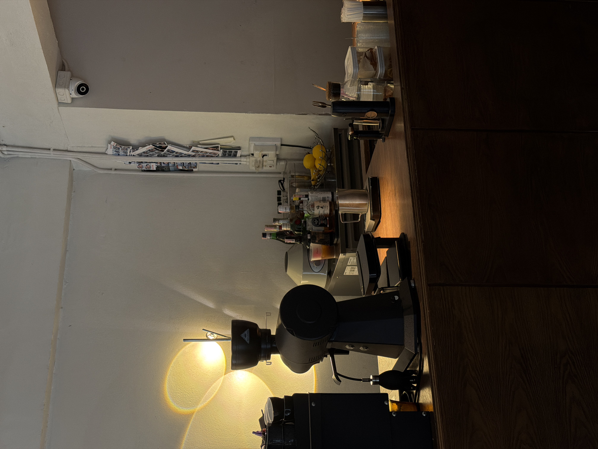 The image depicts a dimly lit interior, likely a coffee shop or bar, with warm lighting casting prominent shadows and creating a sunbeam effect on the wall. In the foreground, a dark wooden counter is laden with various objects. Prominently featured is a large black coffee grinder. To its left, a black rectangular appliance sits next to a small amber glass. To the right of the coffee grinder, there is a stainless steel measuring cup, a small black device with a brass accent, a glass container with liquid and a stopper, and a metal container filled with white paper sleeves. Further to the right, a glass bowl holds several yellow lemons. Behind the counter, bottles of alcohol are visible on shelves, along with a blender or food processor. On the wall above, a security camera is mounted in a white housing. A rack of photographs or postcards is attached to the wall near some electrical conduits. The overall impression is one of a functional space, potentially after closing hours or during a quiet period. The specific location in Chiang Mai City Municipality, Thailand, cannot be determined from the visual information provided.
