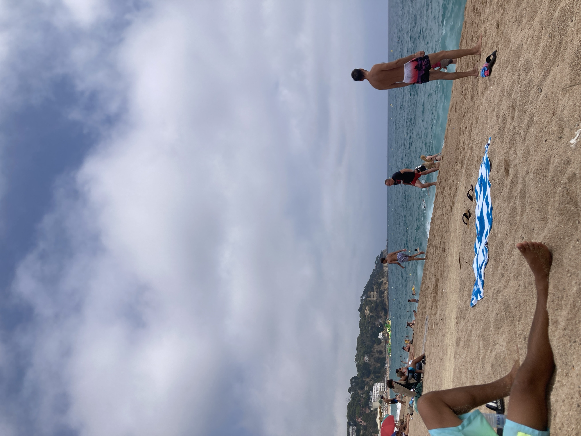 This is a full-frame, eye-level, natural light, outdoor medium shot of a crowded beach in Lloret de Mar, Spain, on a cloudy day. The foreground is dominated by the sandy beach, with a person's legs visible in the bottom left, wearing turquoise and yellow swim shorts. A blue and white striped towel is spread on the sand in the mid-ground, with a pair of black flip-flops next to it. To the right, a man wearing colorful swim trunks stands facing away from the camera, looking out at the ocean. Further back, people are visible wading in the turquoise water, and a large inflatable water park is situated offshore. The background shows a tree-covered hillside with buildings nestled into the slopes, characteristic of the coastal landscape of Lloret de Mar. The sky is overcast with dramatic cloud formations.
