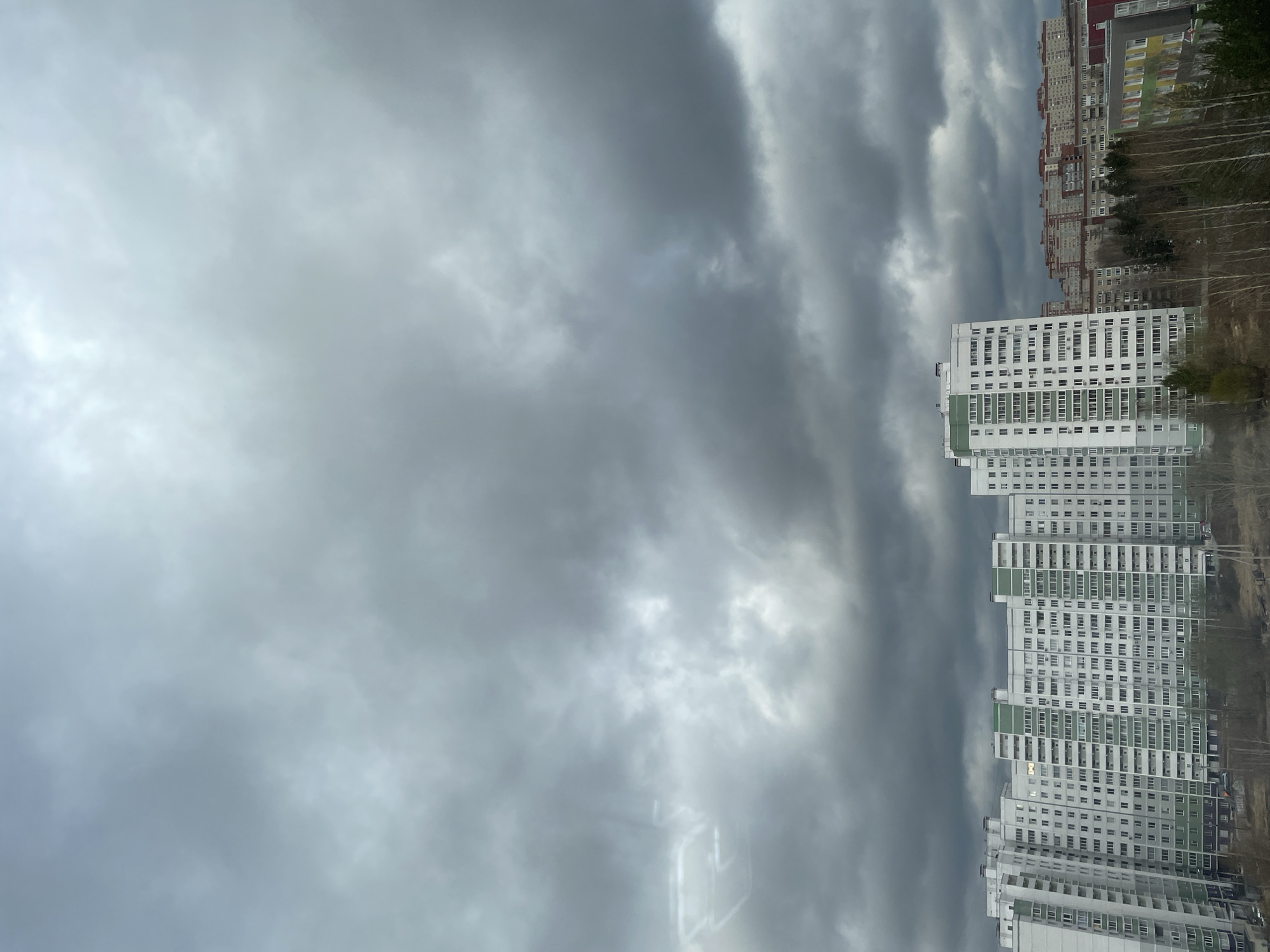 The image captures an urban residential scene dominated by multi-story apartment buildings under a heavily overcast sky.
In the foreground, on the right side of the frame, stands a large, white high-rise apartment building with several bands of light green along its facade. Numerous uniformly sized, rectangular windows are visible across its many floors. Behind this, another similar white and green building can be seen. Further into the background, a sprawling apartment complex features sections with varying facade colors, including red, yellow, and grey, characteristic of post-Soviet era architecture. Bare trees and sparse foliage are visible among and behind the buildings, suggesting a late autumn, winter, or early spring season.
The sky occupies a significant portion of the image, especially the left side and above the buildings. It is filled with thick, dark grey clouds, indicating heavily overcast weather. Some lighter patches within the cloud cover suggest variations in cloud density. The overall impression is one of a dim, possibly chilly, daytime setting.
There are no people or specific activities visible in the static scene. No discernible text is present. The architectural style and dense residential layout are consistent with urban environments in Russia, specifically Izhevsk as per the provided context. The heavy cloud cover creates a somewhat somber or muted atmosphere.