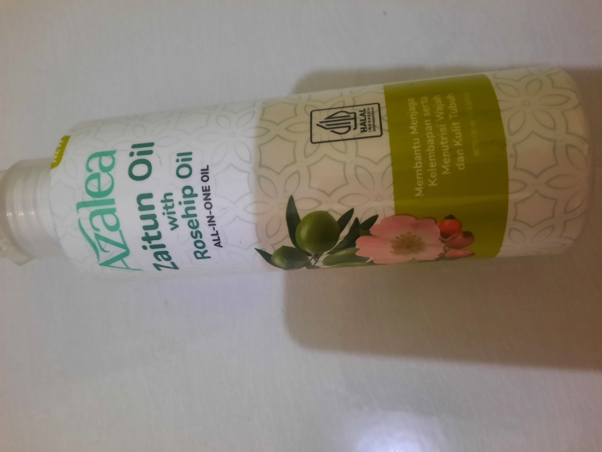 The image shows a bottle of Azalea Zaitun Oil with Rosehip Oil. The bottle is white with green and pink lettering and a floral pattern. A graphic of olives and a pink flower is visible on the side of the bottle. A "Halal Indonesia" logo is also present. Text on the bottle reads "Membantu Menjaga Kelembapan serta Menutrisi Wajah dan Kulit Tubuh" which translates to "Helps Maintain Moisture and Nourish Face and Body Skin". The bottle appears to be on a white surface, possibly a table, with a shadow cast to the left. There is no indication of the city of Lhokseumawe, Indonesia in the image.