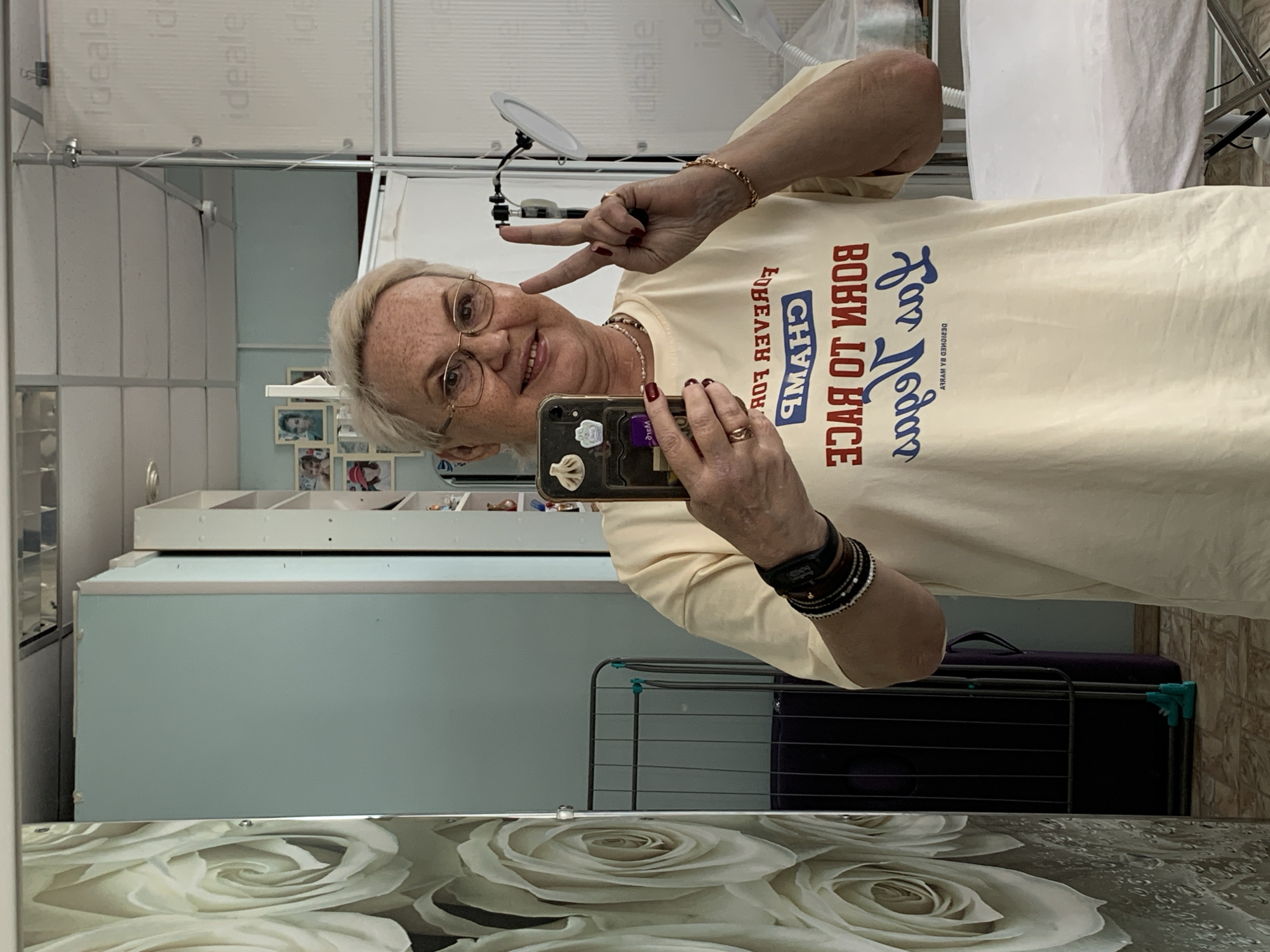 An older woman with short, light-colored hair and glasses takes a mirror selfie in an indoor setting. She is smiling cheerfully and makes a peace sign with her right hand. Her nails are painted dark red, and she wears multiple rings and bracelets on both wrists. She holds a smartphone, likely an iPhone with a clear case and small decorative charms, in her left hand.
She is wearing a light yellow t-shirt. The text on her shirt, visible in reflection, reads (when corrected for the mirror): "For Loony" in blue cursive, "BORN TO RACE" in red block letters, and "ВОИН" (Voin, meaning "Warrior" in Russian) in white letters within a blue rectangular box. Below this, smaller text reads "DESIGNED BY MYREV".
The background, seen through the mirror reflection, features light blue walls and white furniture or shelving units. Several framed photographs are visible on one of the shelves. To the right, a white bed or table covered with white fabric, and a metal drying rack, are partially visible. A flexible lamp with a circular light is positioned behind her right shoulder. The bottom edge of the mirror is adorned with a decorative pattern of white roses against a dark background. The scene appears to be a room in a home or possibly a salon in Chaikovsky, Russia.