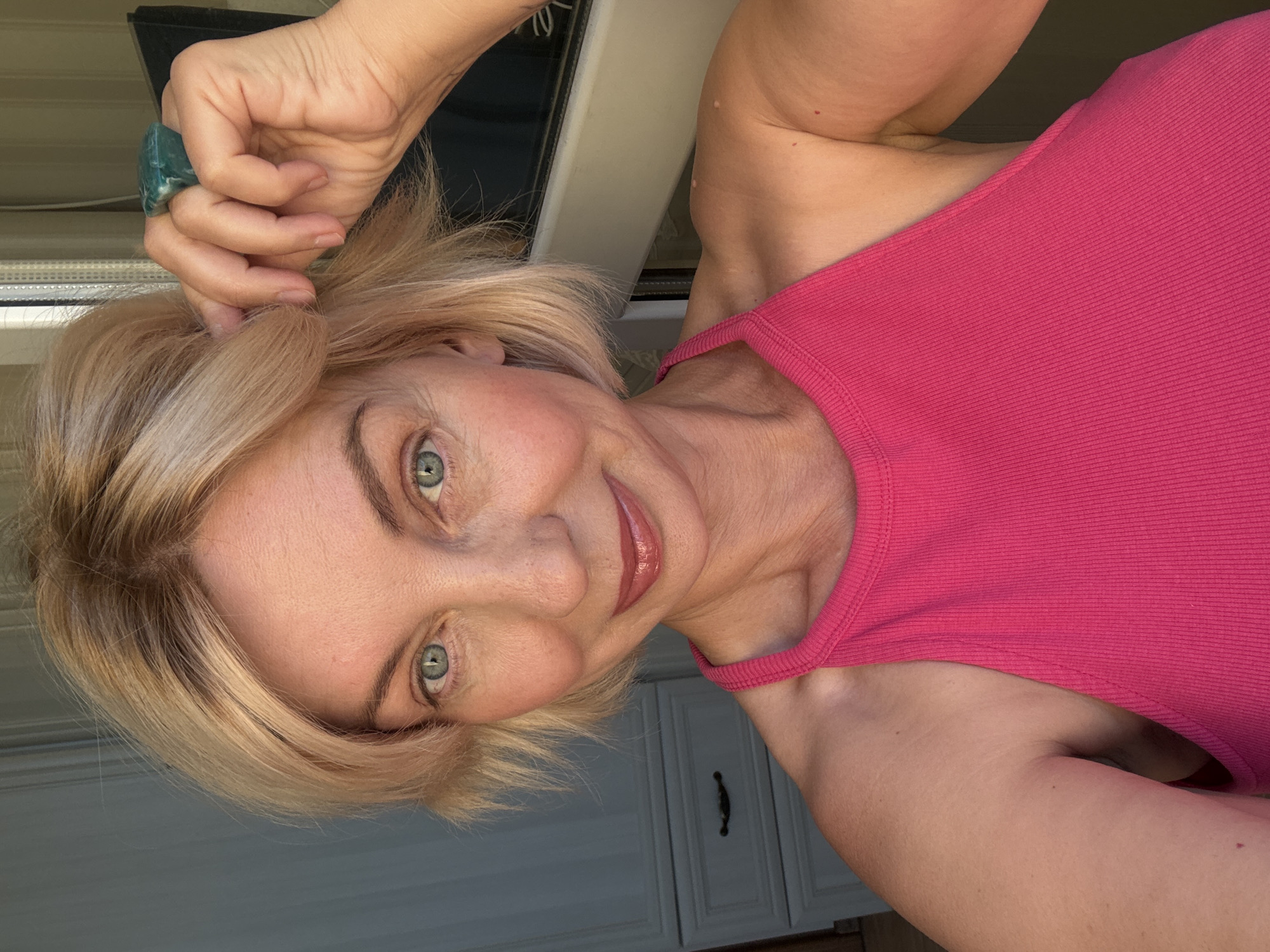 A woman with blonde, short hair and blue eyes is taking a selfie. She is wearing a bright pink tank top and a large, rectangular, green ring on her right index finger. Her right hand is touching her hair near her temple. She is looking directly at the camera with a slight smile. The background appears to be a room with light-colored walls and furniture, possibly a dresser with drawers. The image is taken indoors, with natural light coming from the right side of the frame. No specific landmarks of Sochi, Russia are visible.