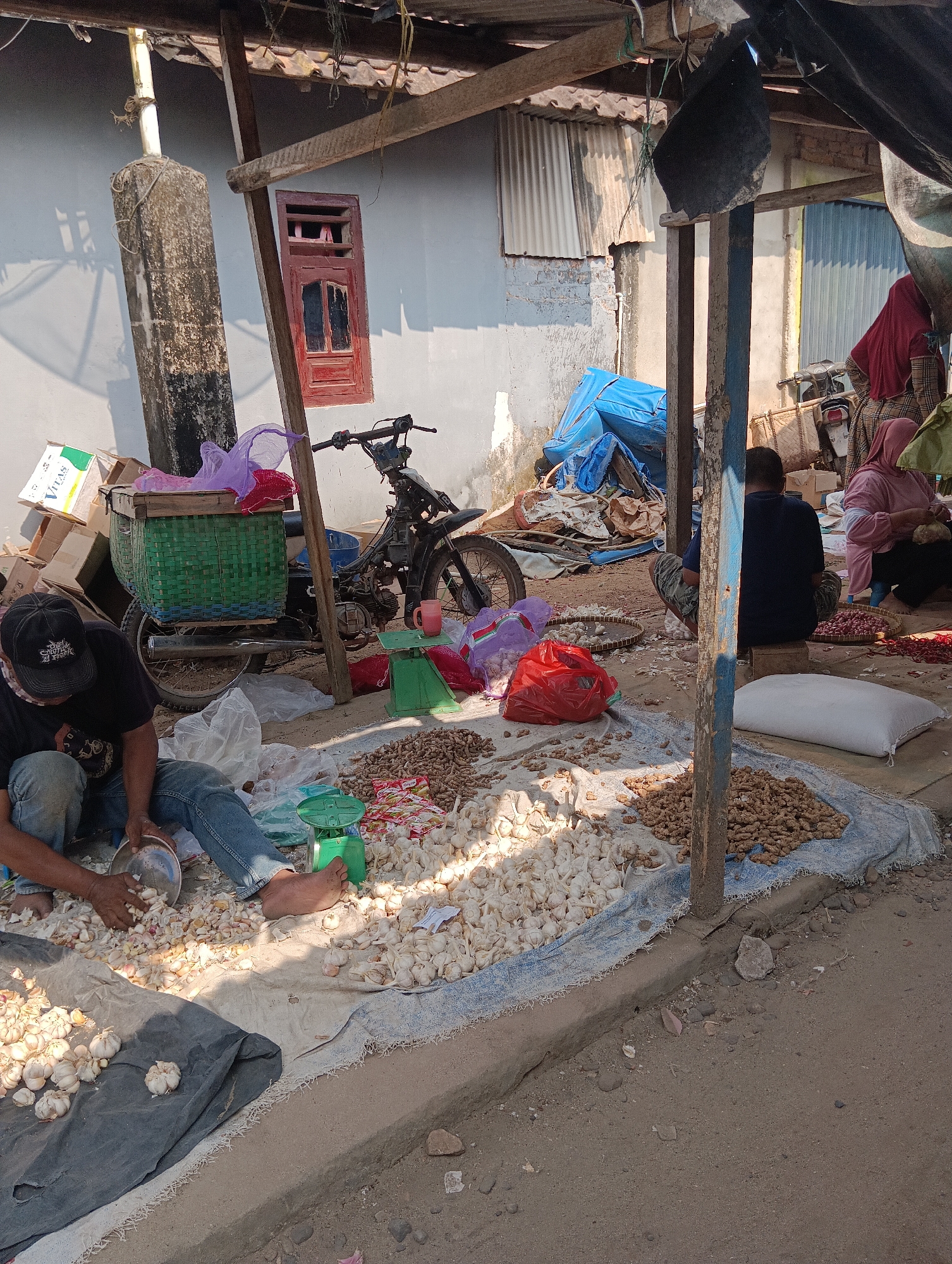 In Binakarsa, Indonesia, a street vendor, a male wearing a black cap, black t-shirt, and blue jeans, crouches on the ground next to a tarp piled with garlic and other produce. He is sorting garlic into a metal bowl. Another person, partially visible, sits on a stool further back, facing away from the camera, near a motorcycle and a scale. To the right, a woman in a pink hijab and patterned clothing is also engaged in sorting produce. The scene is an outdoor market stall, with a makeshift roof supported by wooden poles. Various goods, including sacks of produce, baskets, and boxes, are visible in the background. The ground is dirt and concrete, with some debris. The lighting suggests it is daytime.