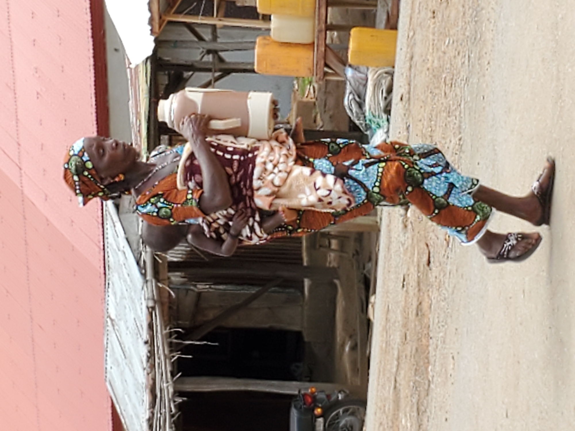 In Mararaba, Nigeria, an adult woman, wearing an intricately patterned headscarf and colorful clothing, carries a large, light-colored jug in her arms. A child is wrapped in patterned cloth and carried on her back. The woman walks on a concrete surface alongside a weathered building with a partially visible red-painted roof and an open structure revealing a motorcycle parked beneath. Multiple yellow containers are stacked behind the woman.