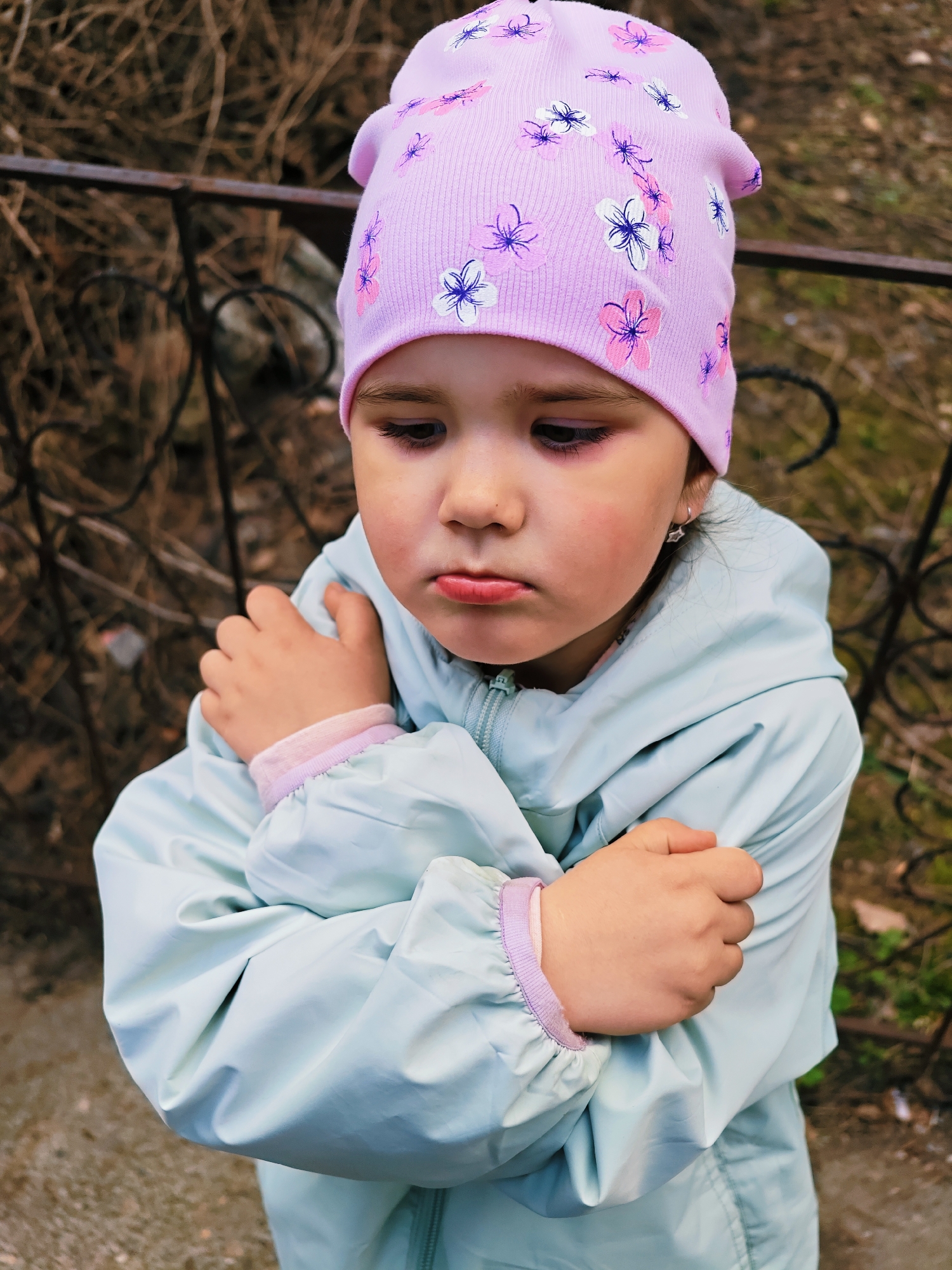 A young girl, standing outdoors in Chaikovsky, Russia, is seen from an elevated perspective. She wears a light blue jacket and a lilac-colored hat with a floral pattern. She crosses her arms tightly across her chest, and her facial expression suggests discontent or cold. In the background, there is a wrought-iron fence and bare branches of shrubs or trees, which, combined with the girl's attire and posture, indicate a cool or transitional season. The ground appears wet or damp, adding to the potentially chilly environment. The photograph captures a moment of apparent discomfort or unhappiness of the child.