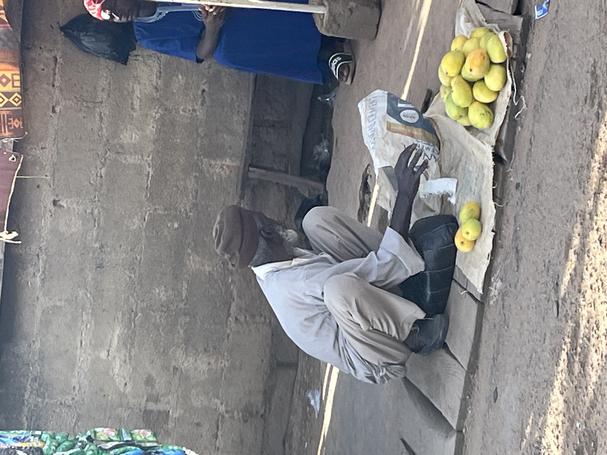 In Gura Top, Nigeria, a man in a brown head covering and a light-colored long-sleeved shirt and pants squats on wooden planks. He is reaching towards a bag of cement. To his right, a pile of yellow mangoes rests on a light-colored cloth. A second person, wearing a blue robe and a red head covering, sits in the background to the right, partially obscured by a dark bag hanging on a rough-textured wall. The environment appears to be an outdoor market setting with a dirt ground and a textured concrete wall.