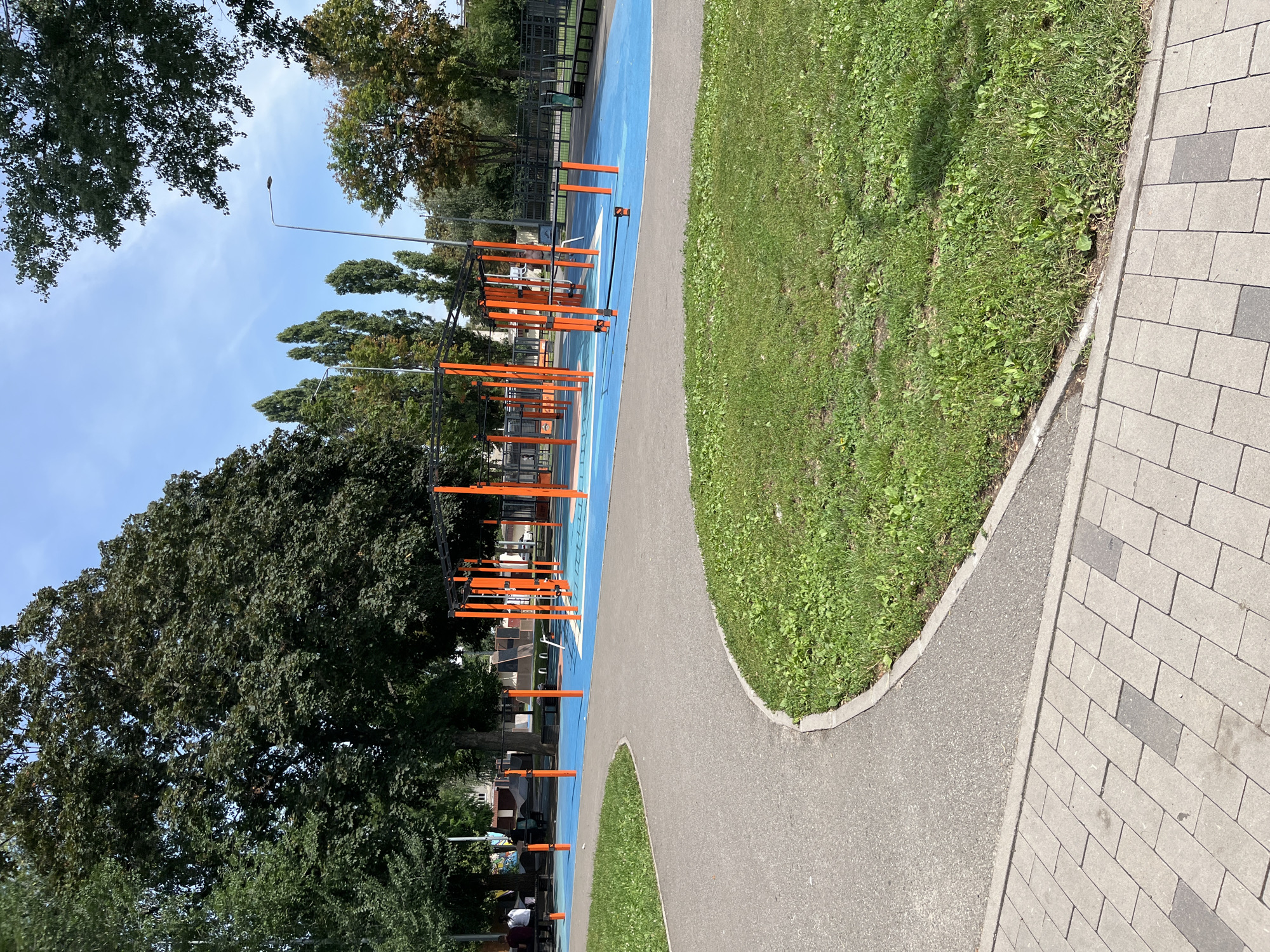An outdoor public park in Kharkiv, Ukraine, features an urban fitness area with a blue rubberized surface. The area is equipped with orange metal structures, including horizontal bars and parallel bars, designed for exercise. In the background, mature trees with dark green foliage form a canopy, and buildings are visible beyond the trees. The foreground shows a grey asphalt pathway curving around a patch of green grass, bordered by light grey brick paving. The sky above is a clear blue with scattered white clouds.