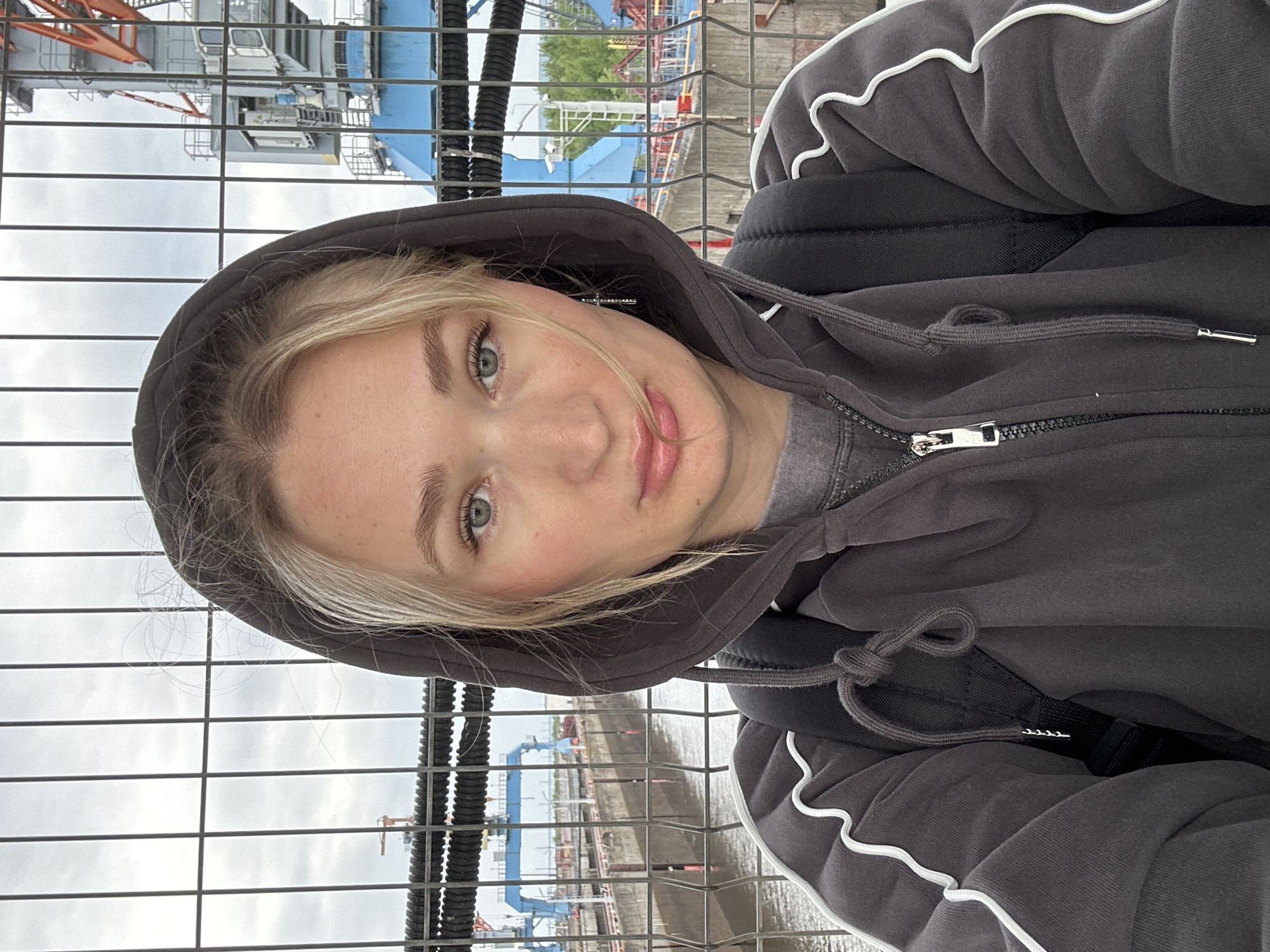 A young blonde woman wearing a dark grey hooded sweatshirt and a black backpack takes a selfie in front of a wire fence. Behind her, a body of water with docked barges and industrial cranes is visible. The sky is overcast. The location is likely a port or industrial area in Chaikovsky, Russia, given the presence of large cranes and industrial structures commonly found in such environments.