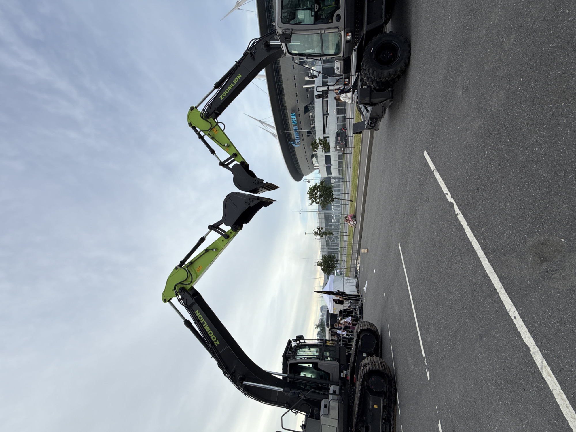 Two Zoomlion excavators are positioned facing each other on an asphalt road in front of the Gazprom Arena stadium in Saint Petersburg, Russia. The stadium, with its distinctive white, sail-like roof structures and the "GAZPROM ARENA" logo visible, serves as the backdrop. The excavator on the left is a tracked model, while the one on the right is wheeled. Both excavators have their booms raised and buckets extended, creating a V-shape in the center of the frame. Several white tents and some people are visible near the left excavator, suggesting a potential event or display. In the background, beyond the stadium, is a slightly overcast sky with wispy clouds. The road has visible white lane markings.
