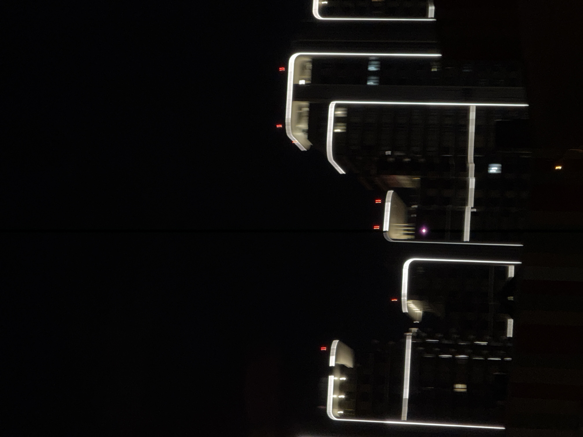 The image, taken at night in Khimki, Russia, captures a striking architectural view of a modern building defined by its linear illumination. The structure is silhouetted against a completely dark background, emphasizing the bright, glowing white light strips that trace its contours. These lights outline various geometric sections of the building, including horizontal ledges or floor levels and vertical pillars, creating a sleek and contemporary aesthetic.
Interspersed along these primary white lights are smaller, distinctive red lights, often appearing in pairs of wavy or chevron shapes. These red accents punctuate the design, possibly serving as decorative elements or functional markers. Faint reflections of internal lighting are visible through some windows or glass panels, hinting at the building's interior.
A prominent feature of the composition is the lower half of the image, which presents a near-perfect, mirrored reflection of the illuminated building above. This suggests the presence of a highly reflective surface, such as still water or a polished dark ground, creating a powerful sense of symmetry and enhancing the dramatic visual effect. The scene evokes a sense of modern urban elegance and architectural design. No people or discernible text are visible.