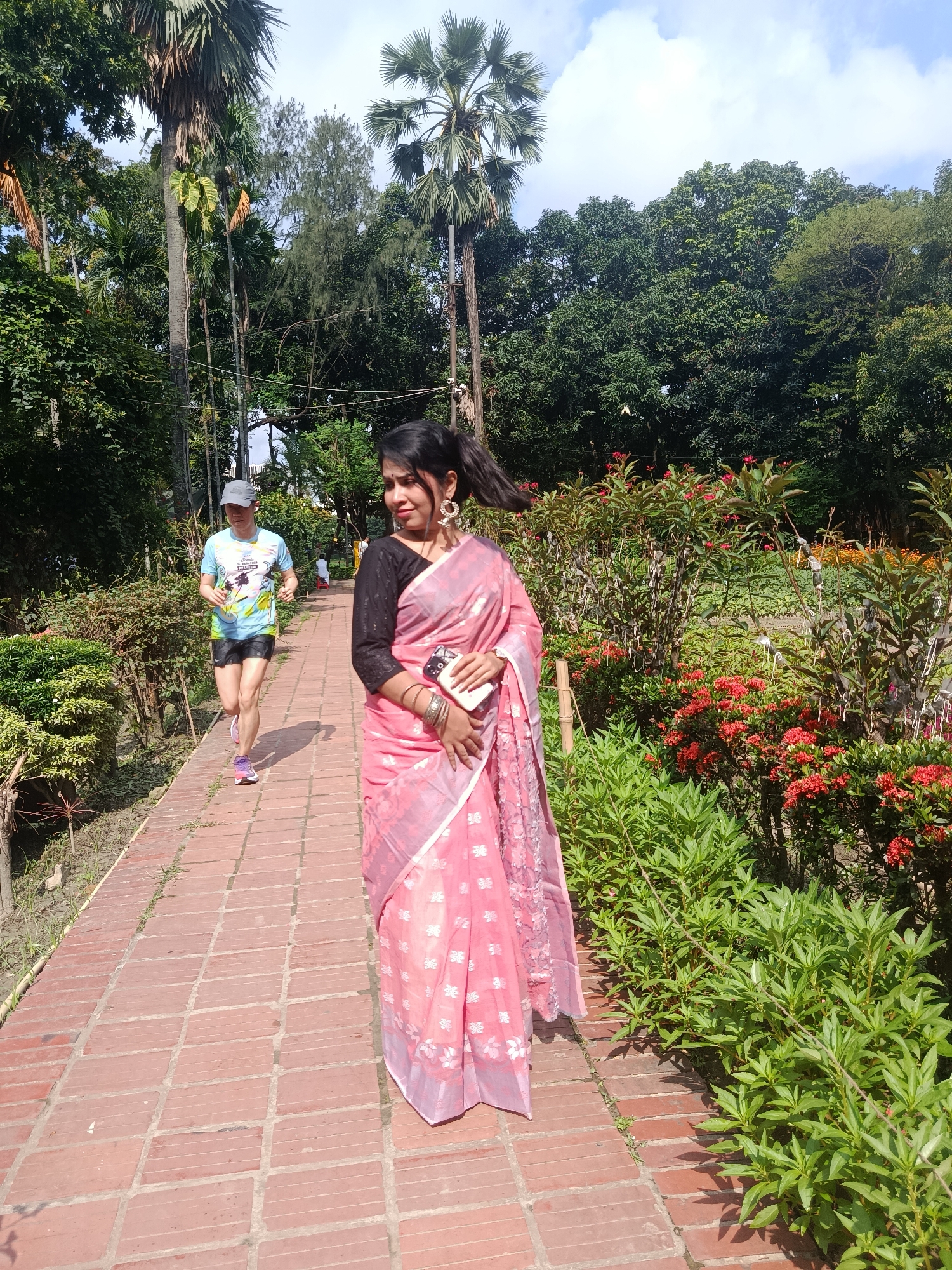 The image shows a woman in a pink sari and black long-sleeved blouse standing on a brick pathway in a garden. She has dark hair, earrings, and is holding a cell phone and wearing a wristwatch. A man is jogging on the same path, wearing a light blue T-shirt, gray cap, and dark shorts. Red flowers and green bushes border the path on the right side. In the background are various trees. The location is likely in the city of Dhaka, Bangladesh.