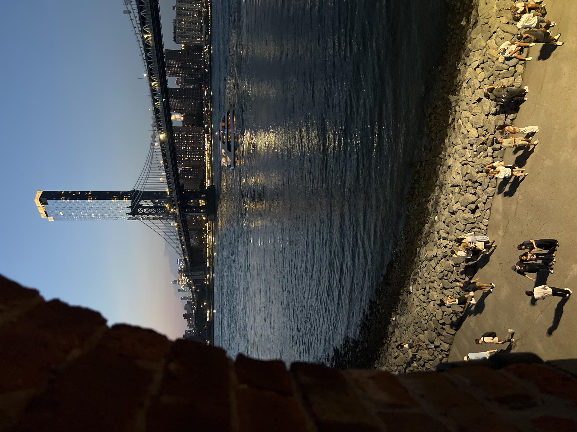 The image captures a dusk scene along the East River in New York City, with the Manhattan Bridge dominating the mid-ground and a modern skyscraper to its left. The perspective is from a brick structure on the left, creating a partial frame.
In the foreground, a pathway lines the riverbank, which is fortified with large, irregular stones. Several groups of people are seen strolling and conversing on this pathway. Their attire suggests a casual, evening outing. Some individuals are standing, while others are walking. Shadows from an unseen light source are cast long across the pathway.
The East River is a dark expanse of water, reflecting the lights of the city and a pleasure boat navigating the river. The boat, with illuminated windows, is positioned to the right of the bridge's central span.
Beyond the river, the Manhattan Bridge stretches across, its lights illuminated. The New York City skyline, featuring numerous buildings with lit windows, is visible in the distance, contributing to the urban landscape. The sky transitions from a deep blue to lighter hues near the horizon, indicative of twilight. The skyscraper to the left of the bridge is notably taller than the surrounding buildings, its glass facade reflecting the sky and city lights.