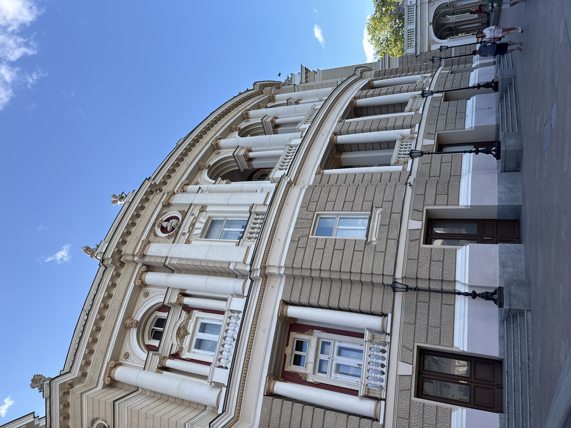 The image shows the exterior of a grand, ornate building, likely an opera house or theater, in Odesa, Ukraine. The architecture is classical, with prominent columns, arched windows, and decorative stonework. The building is bathed in bright sunlight, indicating it's daytime. The sky is a clear, vibrant blue with a few wispy clouds, suggesting pleasant weather.
In the foreground, on the right side of the image, several people are walking up a wide, stone staircase towards the entrance of the building. They appear to be casually dressed tourists or visitors. There are also decorative lampposts lining the staircase. The building's facade is a mix of light-colored stone and textured grey panels, with accents of deep red around some of the windows. The overall impression is one of historical grandeur and architectural beauty. The low angle of the shot emphasizes the impressive scale of the building.