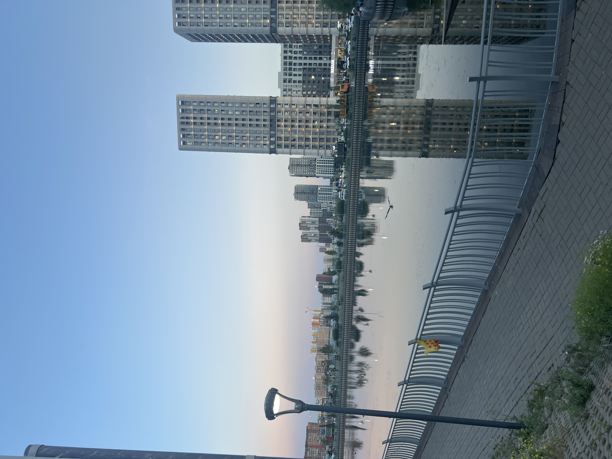 This is a photograph of the city of Astana, Kazakhstan, likely taken in the evening or early morning. The scene is dominated by a body of water, possibly a canal or river, reflecting the surrounding urban landscape and the sky. On the right side of the image, several modern, multi-story apartment buildings with numerous windows are visible, their reflections clearly discernible in the water. In the middle ground, a row of smaller buildings and trees lines the opposite bank of the water. A pedestrian walkway with a metal railing runs along the foreground, leading the viewer's eye into the scene. A streetlamp stands on the walkway, and a yellow plastic bag is hanging from the railing. The sky transitions from a pale blue at the top to a soft pink and orange hue near the horizon, indicating either dawn or dusk. A bird is in flight over the water in the middle of the frame. The overall atmosphere is serene and urban.