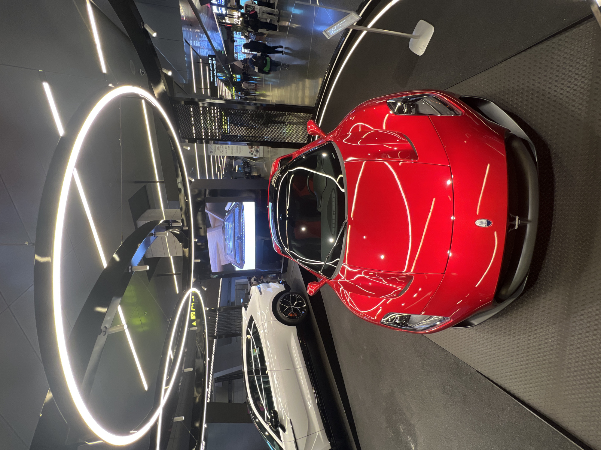 The image captures a vibrant red supercar, identified as a Maserati MC20 by its distinctive design and front trident emblem, prominently displayed in a sleek, modern indoor setting. The car is positioned diagonally on a dark, textured, checkered plate platform. Behind it, a white Maserati, possibly an SUV or sedan, is partially visible.
The environment appears to be a high-end showroom or a luxury exhibition area within a large public space, potentially an airport or a mall, suggested by the blurred figures of people walking in the background and signs indicating directions (including "EXIT") in both Arabic and English script.
The scene is brightly illuminated by striking circular and linear LED light fixtures that cast brilliant reflections across the glossy surfaces of the cars, highlighting their contours. The floor surrounding the display area is a polished, reflective dark surface. The overall atmosphere is one of modern luxury and sophistication. The time of day is not discernible as it is an indoor setting with strong artificial lighting.