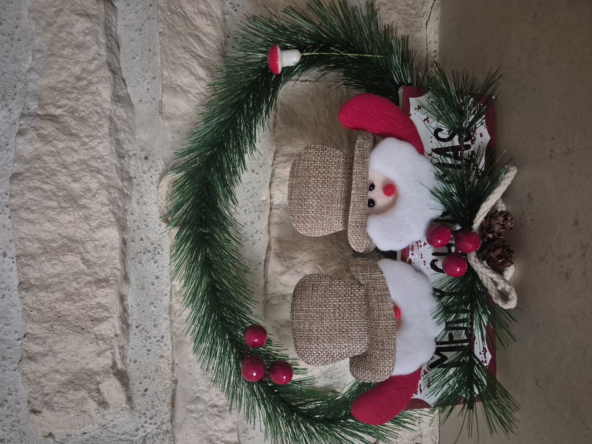 A Christmas wreath, adorned with festive decorations, is depicted against a textured, light-colored wall. The main subject is a circular green artificial pine wreath, oriented horizontally despite the image being vertically composed.
Within the wreath, two decorative Santa Claus figures are prominent. Each Santa has a fluffy white beard, a small red nose, and black eyes. Their hats are red, and their bodies or lower hat portions are made of a burlap-like textured fabric in a light brown color.
The wreath itself is embellished with several items:
- On the upper left side, a small red-capped mushroom with a white stem is attached to the pine needles.
- On the lower left side, three shiny red berries are clustered together.
- On the right side, between the two Santa figures, another cluster of three red berries is visible.
- Below these berries, a small pine cone and a loop of twisted rope are integrated into the design.
Behind the Santa figures and extending to the right, there is a red and white sign or board. Partial text is visible on this sign. The clearest legible word is "MERRY" on the lower right part of the sign. Other fragmented letters like "S" and possibly "ME" can also be discerned, suggesting a common Christmas greeting like "Merry Christmas" or "Merry and Bright" on a distressed white background with red patterns.
The background consists of an unevenly textured wall, appearing to be made of light beige or grey stone, concrete, or a similar material, providing a neutral backdrop for the colorful holiday decoration.