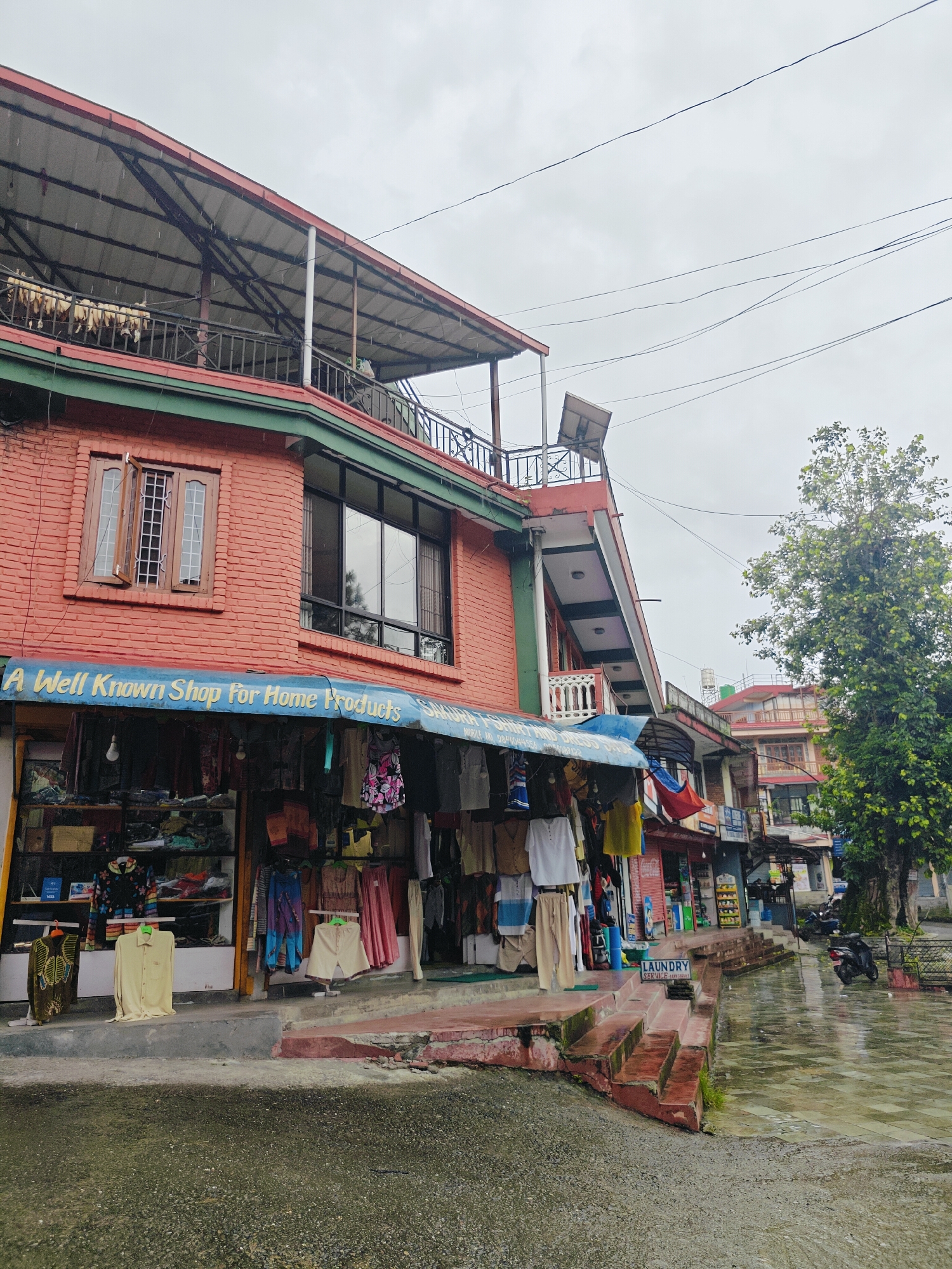 A retail establishment in Pokhara, Nepal, displays various home products and clothing items. The shop, constructed from red brick, features a prominent blue awning with white lettering that reads "A Well Known Shop For Home Products." Merchandise is displayed both inside the store and hung outside. The street in front of the shop is wet, suggesting recent rainfall, and a tree with lush green foliage stands to the right. A sign for "LAUNDRY SERVICE" is visible on a set of red stairs leading up to the shop entrance. Several motorcycles are parked further down the street. The sky is overcast.