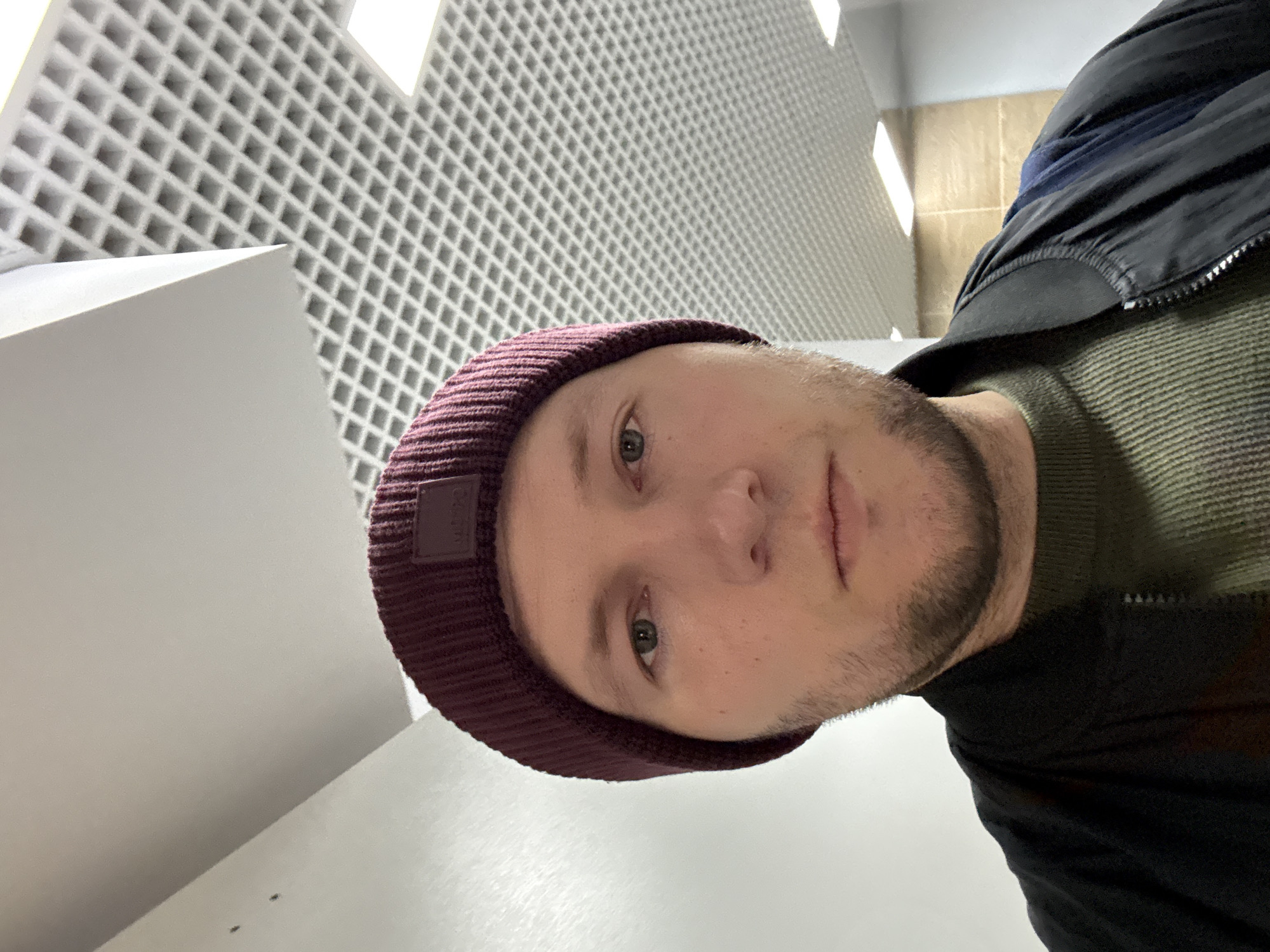 A selfie-style photograph shows a man with a burgundy beanie indoors. The man has light skin, short, light-brown hair, and a light beard. He is wearing a green sweater and a dark-colored jacket. The background features a white, gridded ceiling and a white wall. The location appears to be an interior space, possibly a commercial or public building. The photo provides no information confirming the location to be in Krasnoyarsk, Russia.