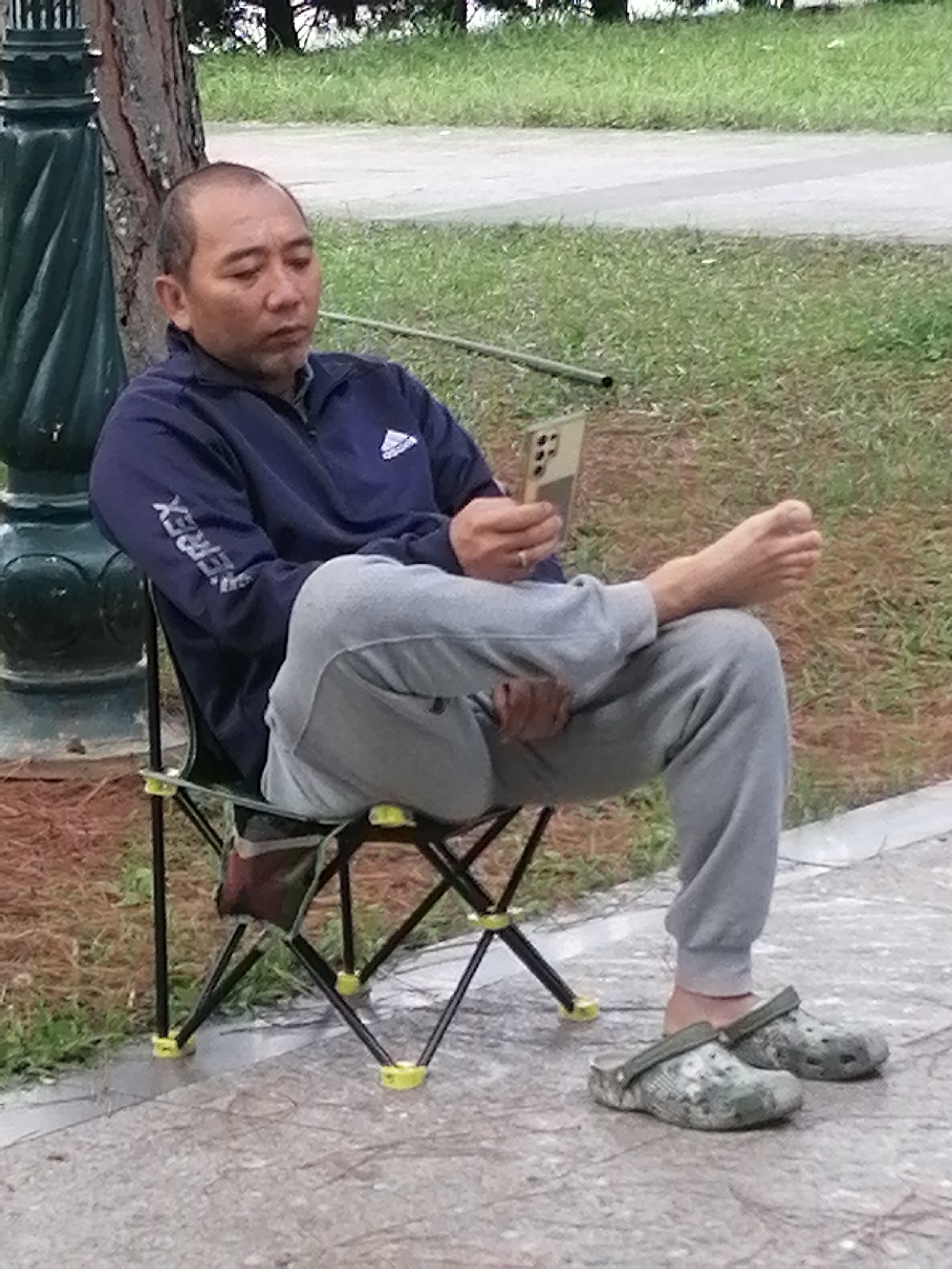 A middle-aged man sits in a portable black folding chair, looking down at his smartphone. He is wearing a navy blue Adidas jacket with "EVEREX" printed on the sleeve, grey sweatpants, and camouflage-patterned Crocs. His left leg is crossed over his right, and his right foot is bare except for his big toe. The man has a serious expression on his face as he focuses on his phone.
The scene is outdoors, with a tree and grass in the background. A paved path is visible in the foreground. The ground appears to be wet, suggesting recent rain or damp conditions, perhaps indicative of the misty, cool weather often experienced in Da Lat. The lighting suggests it is daytime, possibly late afternoon given the soft, diffused light. The overall atmosphere is one of quiet repose, with the man engrossed in his digital device amidst a natural, park-like setting.