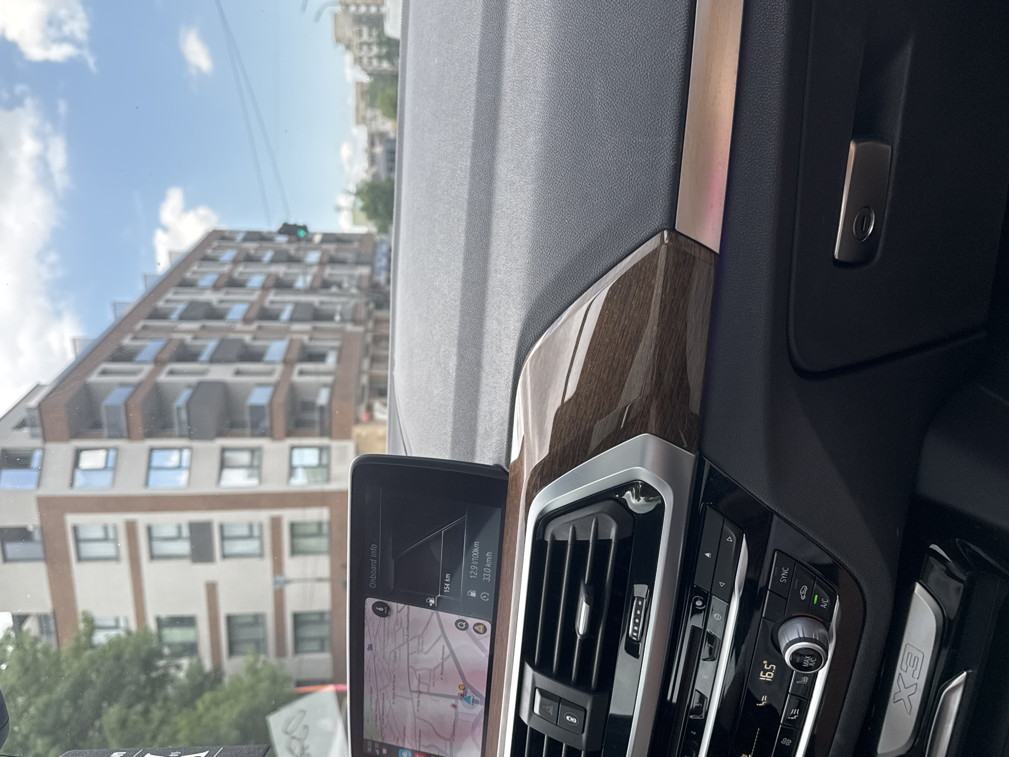 This media file captures the interior of a BMW X3 car, looking out through the windshield at a city environment in Sofia, Bulgaria. The image is oriented vertically (rotated 90 degrees clockwise).
The foreground features the car's dashboard, displaying modern controls and an infotainment screen. On the screen, "Onboard info" is visible, along with car performance data: "154 km" (possibly range), "12.9 l/100km" (fuel consumption), and "33.0 km/h" (average speed). A navigation map is also displayed on the lower part of the screen, showing a street layout. The time "1423" (2:23 PM) is faintly visible at the top left of the screen. To the right of the screen are the car's air vents and climate control panel, which shows a temperature setting of "16.5°" Celsius, along with buttons for "SYNC" and "A/C". The "X3" model branding is visible on the passenger side trim. The dashboard features wood-grain accents and metallic trim, indicating a premium interior.
Outside the car, a multi-story building with numerous windows and balconies is the dominant feature, suggesting a residential or commercial district. The sky is bright with scattered clouds, indicating a daytime setting. A green traffic light is faintly visible above the building in the distance, implying the car might be stopped or moving slowly. Trees and other distant buildings can also be seen. The overall scene depicts a typical urban setting during daylight hours.