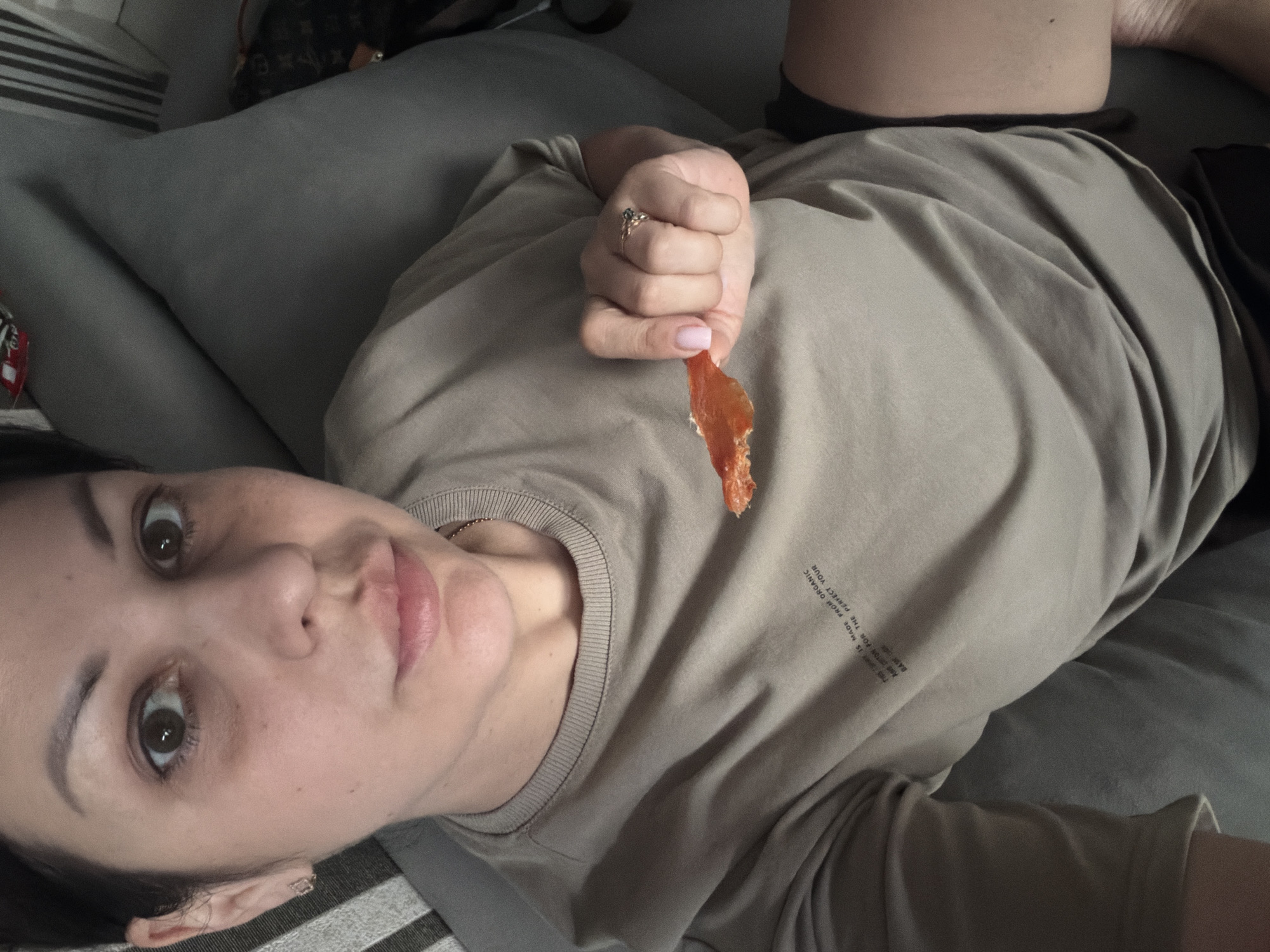 A woman is lying on a gray couch, holding a piece of food in her right hand. She is wearing a tan t-shirt and black shorts. The woman has dark hair pulled back, and she is looking at the camera. The room appears to be dimly lit, with striped cushions on the couch and a bag on the floor to the right. The background is out of focus, but it appears to be an indoor setting. It is not possible to determine the location in the city of Sochi, Russia from the provided image.