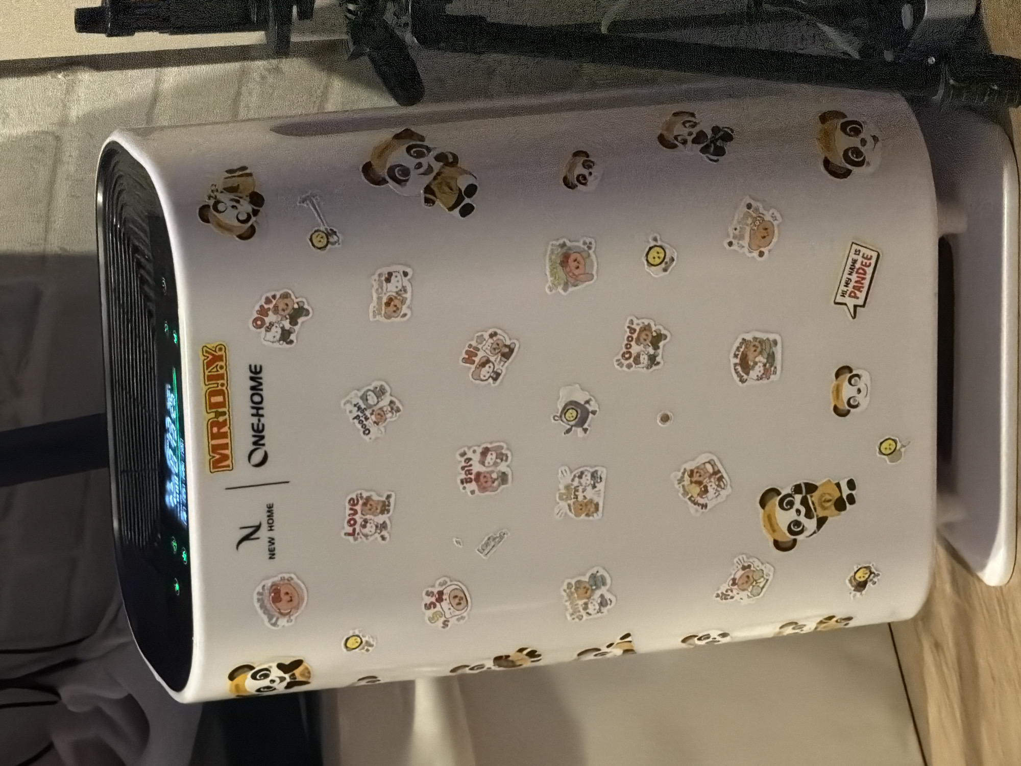 The image shows a white air purifier adorned with numerous stickers, primarily featuring cartoon pandas and bears. The air purifier has a black grill on the left side, with green lights visible through it. A logo on the side reads "MR.DIY ONE-HOME NEW HOME". The stickers display various cute characters in different poses and some with text like "Love", "Good night", and "HI, MY NAME IS PANDEE". The background is blurred, but appears to be a textured wall and a wooden surface, suggesting an indoor setting. The lighting suggests it might be daytime or well-lit indoors. The overall impression is that of a personalized or decorated appliance. There are no people visible in the image. The location context is Bangkok, Thailand.