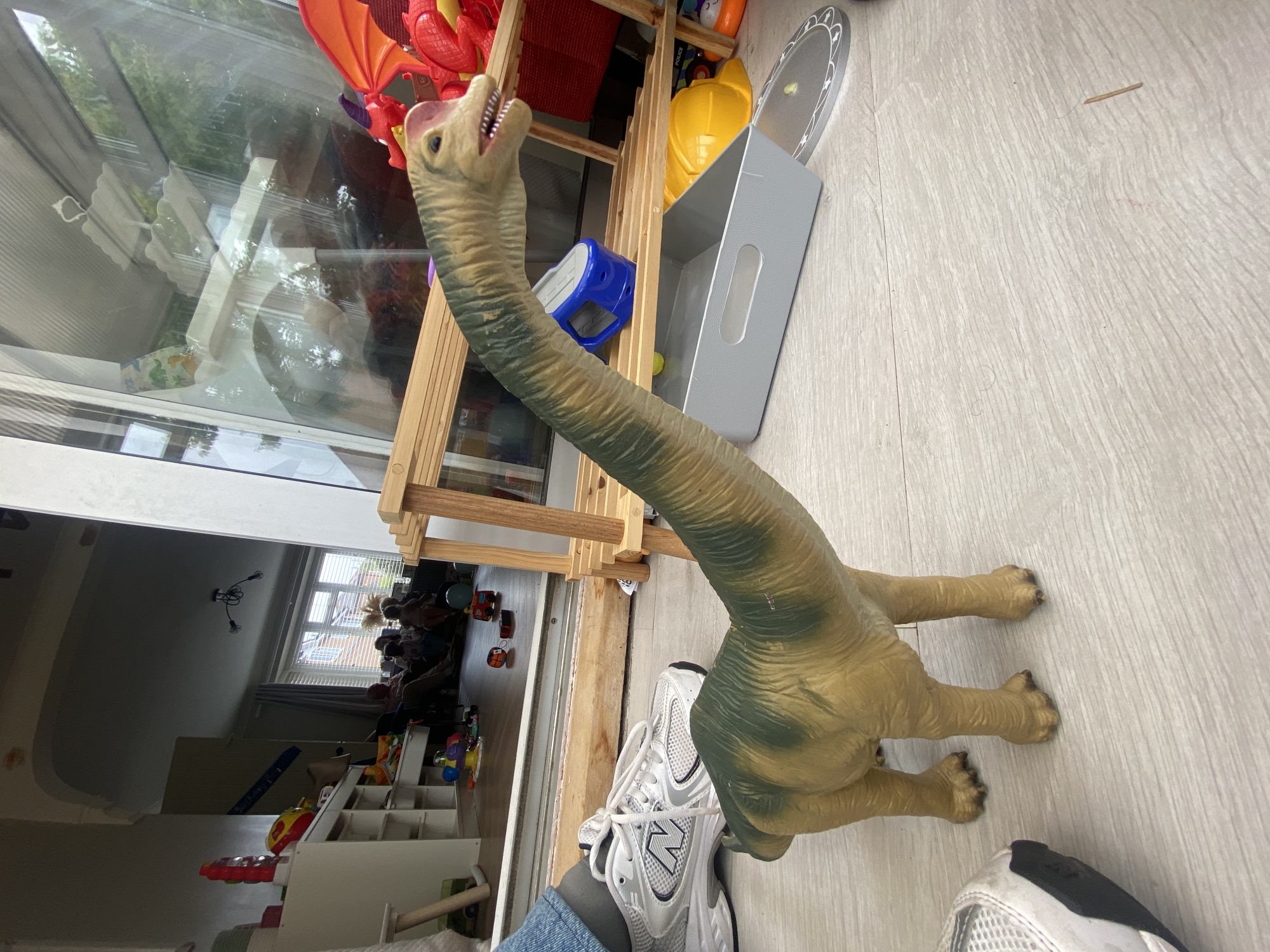This image, captured in Melksham, United Kingdom, displays an indoor scene dominated by a large dinosaur toy. The toy, appearing to be a sauropod (long-necked dinosaur) with a long neck and tail, stands upright on a light-colored wooden plank floor. Its coloration is primarily tan with irregular dark green patches on its back and sides. To the immediate left of the dinosaur’s lower body, a person’s left foot is visible, wearing a white and grey New Balance sneaker with grey socks. The sole of the sneaker is partially visible, indicating a low-angle perspective.
In the mid-ground, a wooden sliding door frame separates the viewer's space from an adjacent room. Through this large opening, another interior space, likely a living room or playroom, is visible. This room features a white shelving unit filled with various colorful toys, including a red and yellow striped ball. Scattered on the light-colored floor of this adjacent room are more children's toys, such as a red toy truck, a multi-colored ball, and a purple and yellow spinning top. Several individuals are seated or kneeling in the background of this room, their features indistinct due to distance and lighting, suggesting an informal gathering. A window with white horizontal blinds is visible at the far end of this room, and a dark, arched architectural feature is present on the upper left wall. A dark light fixture with three visible bulbs hangs from the ceiling.
To the right of the dinosaur toy, a light wooden shelving unit or easel structure stands. Beneath this unit, a grey rectangular storage bin and a round silver tray with star cutouts are placed on the floor. Partially visible behind the wooden shelving, a red dragon toy with prominent orange wings is present. A yellow hard hat and a blue container are also discernible on the floor under the shelving unit. The large glass panel of the sliding door on the right side of the image reflects green foliage, indicative of an outdoor environment, and portions of a white interior ceiling or structure. A small white mobile-like object is also reflected near the top of the glass panel. The overall lighting suggests natural light entering from the right side of the frame. A small wooden stick or splinter is visible on the floor to the right of the dinosaur's foot.
